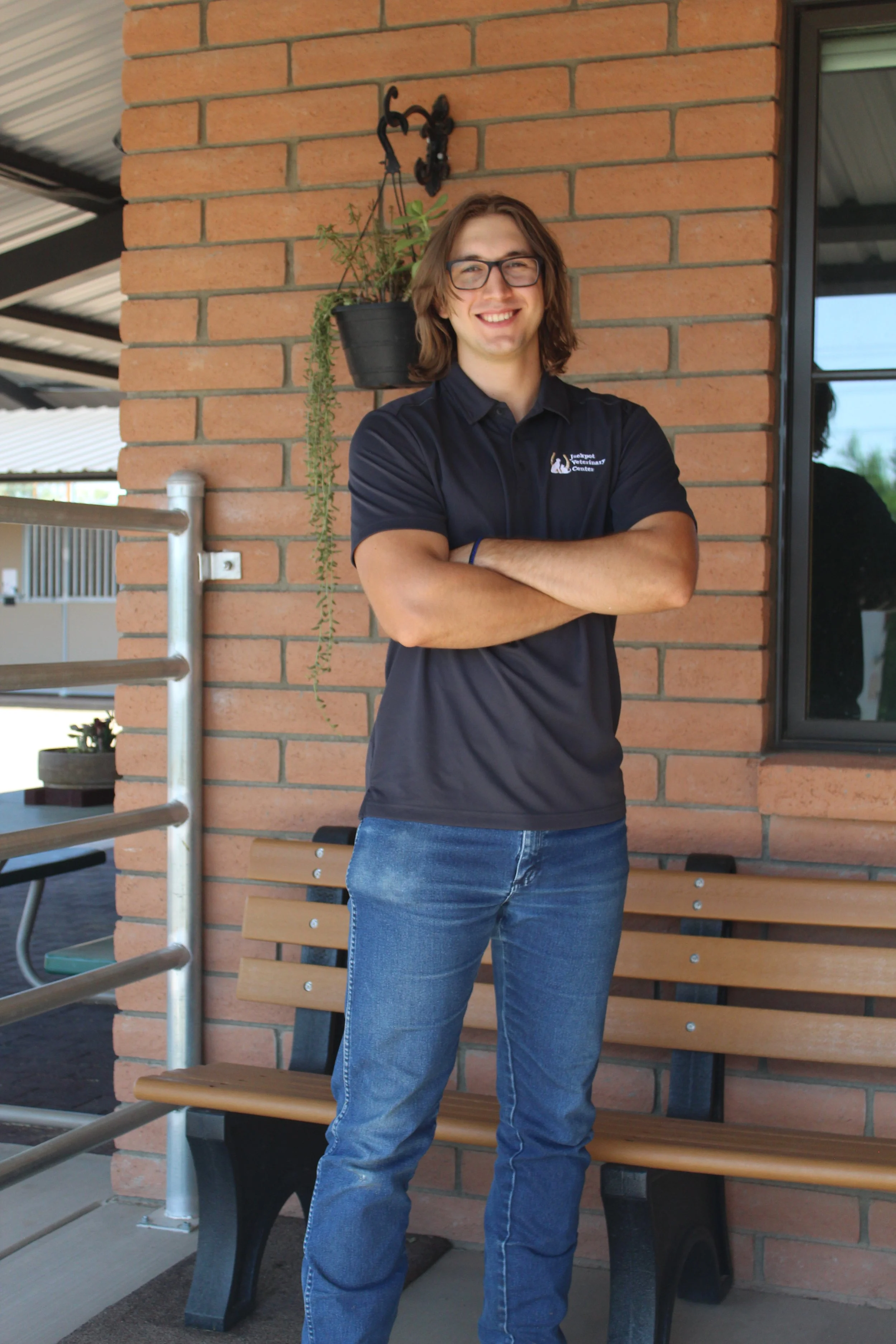 Person in glasses and polo shirt standing with arms crossed, next to a brick wall and a bench, with a potted plant hanging overhead.