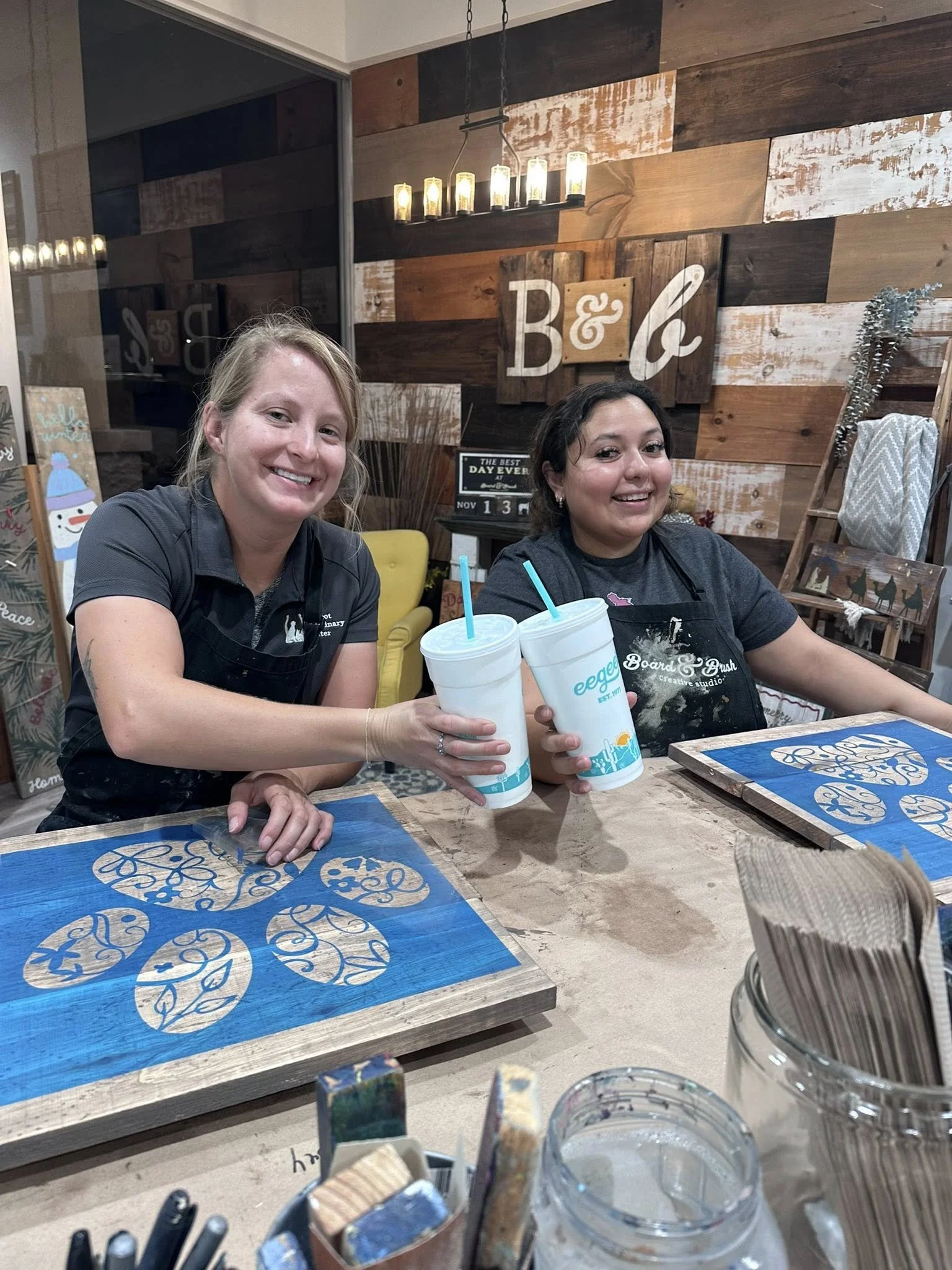 Two women sitting at a table in a decorated room, each holding a large cup with a straw. Behind them is a wooden wall with a sign that reads 'B & b' and various decorations, including a ladder, plants, and art pieces.