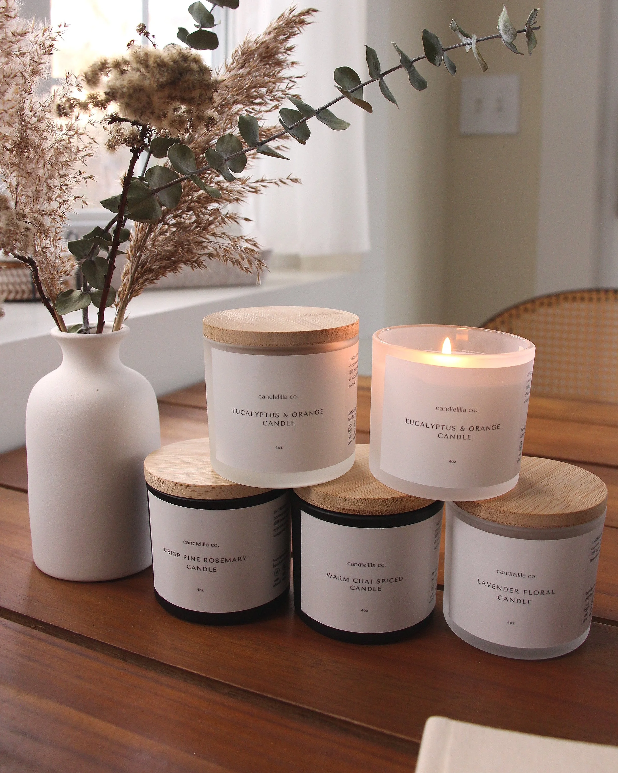 Five scented candles displayed on a wooden table, featuring eucalyptus and orange, crisp pine rosemary, warm chai spiced, and lavender floral scents. One candle is lit, and a vase with dried plants is nearby.