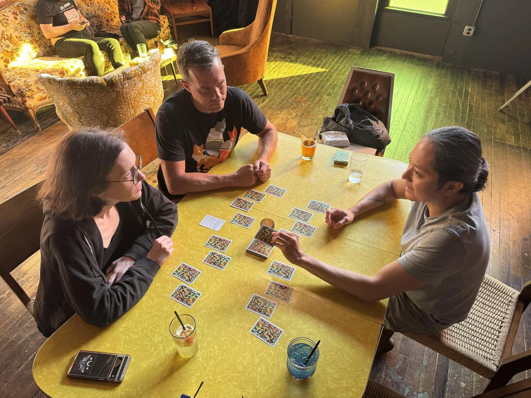 Three people sitting around a yellow table playing a colorful card game. Drinks are on the table, and there are chairs and sofas in the background.