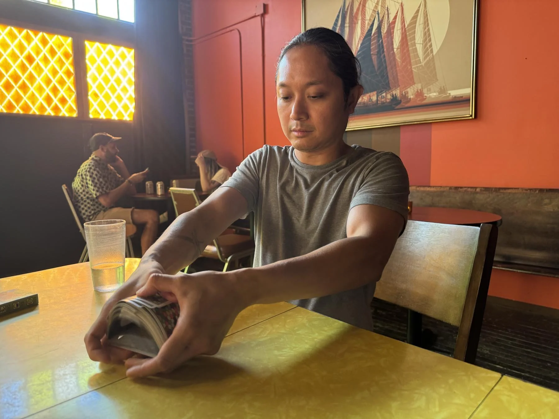 A young man with long dark hair in a ponytail, wearing a gray T-shirt, is sitting at a yellow table in a colorful, well-lit restaurant. He is playing with a deck of cards. Behind him, two people are sitting at another table, one man with a cap and patterned shirt and a woman with long hair, both engaged with their phones. The restaurant features orange walls, a painting of a sailing ship, and patterned window panels.