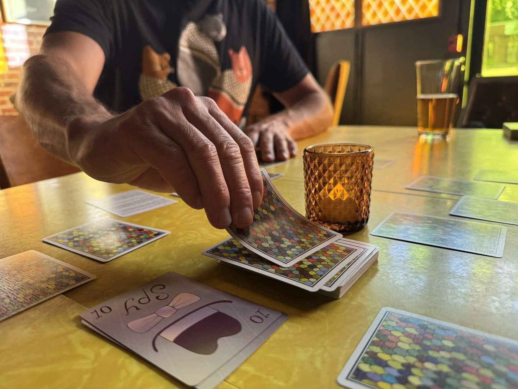 A person playing a card game at a table with amber-colored candles and drinks. The person is holding a deck of playing cards.