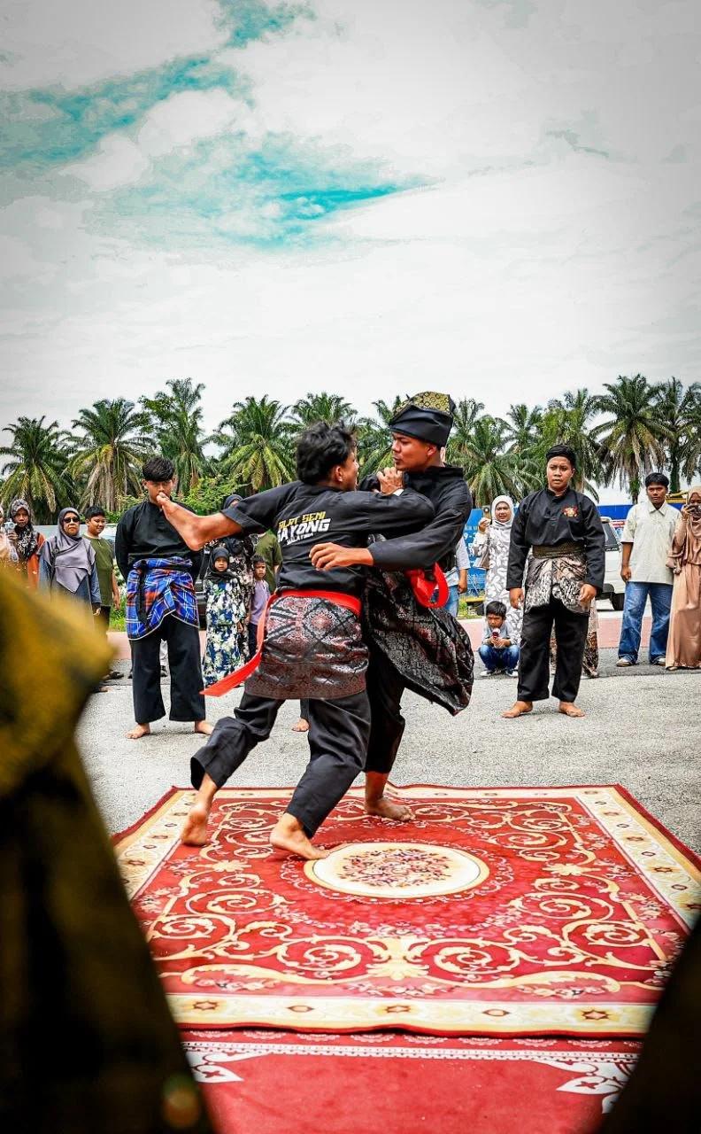 Two men in black martial arts uniforms practicing martial arts on a red rug outdoors, surrounded by onlookers, with palm trees and a cloudy sky in the background.