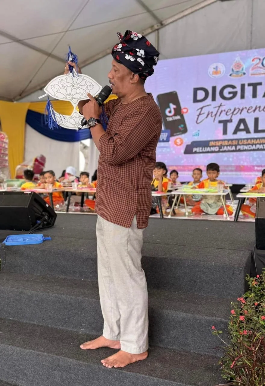 A man wearing traditional attire and a headwrap holding a microphone and a paper cutout, standing barefoot on stage while speaking at a cultural or community event. Several children are seated behind him, and a large screen in the background displays