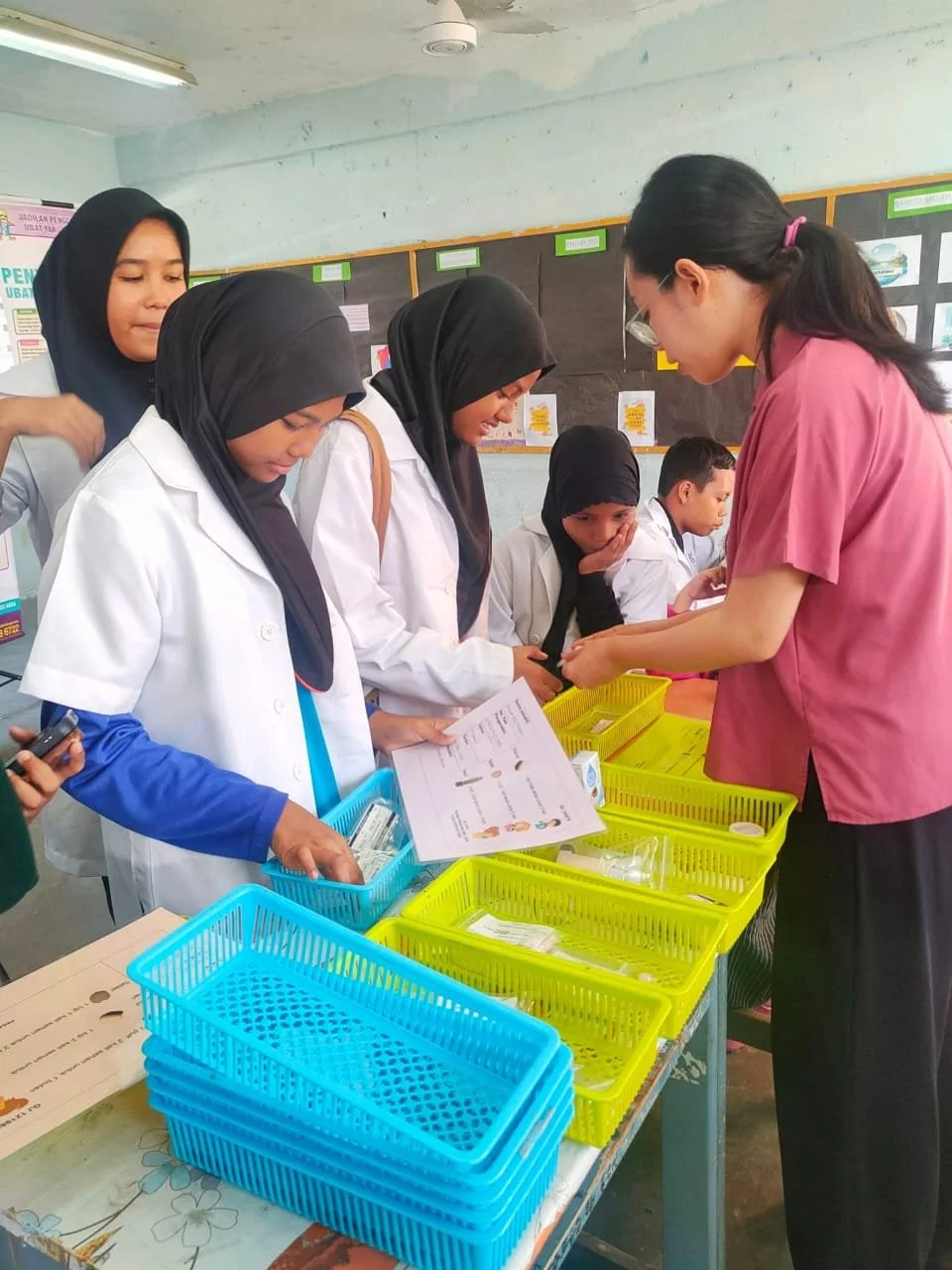 Group of students and a staff member at a health or registration booth, with colorful plastic baskets on the table.