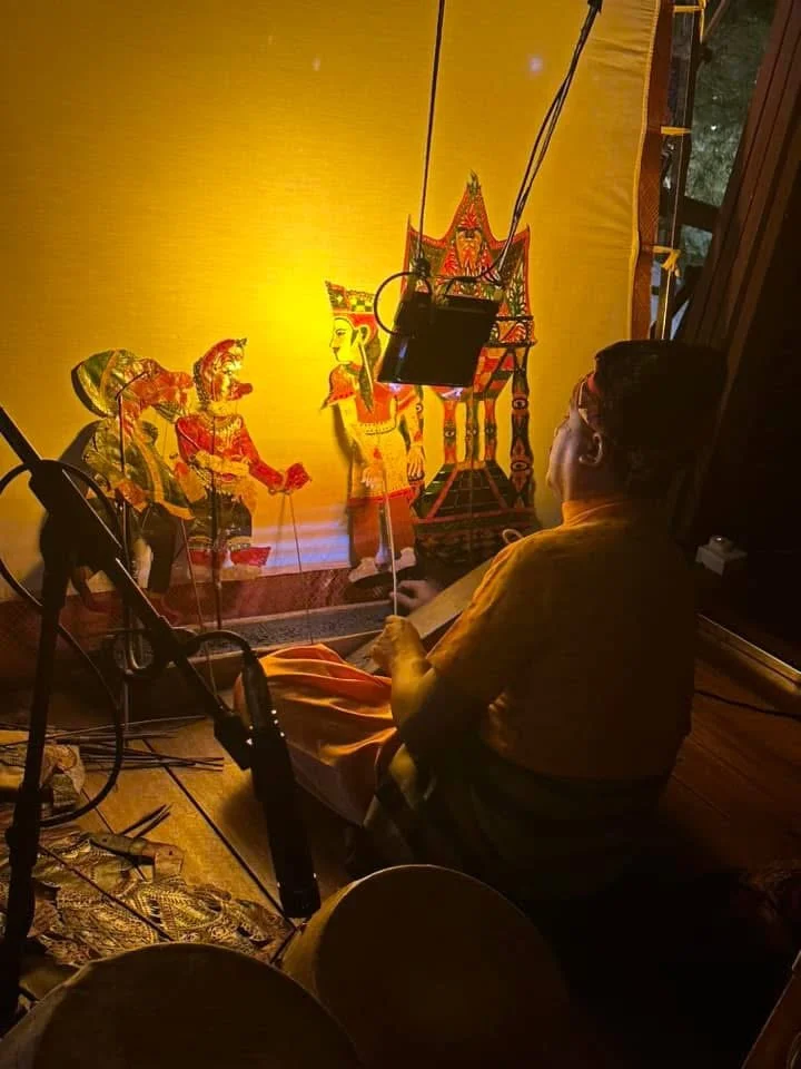 A person sitting on the floor near a traditional shadow puppet, with a yellow background and stage lighting, preparing for a shadow puppet performance.