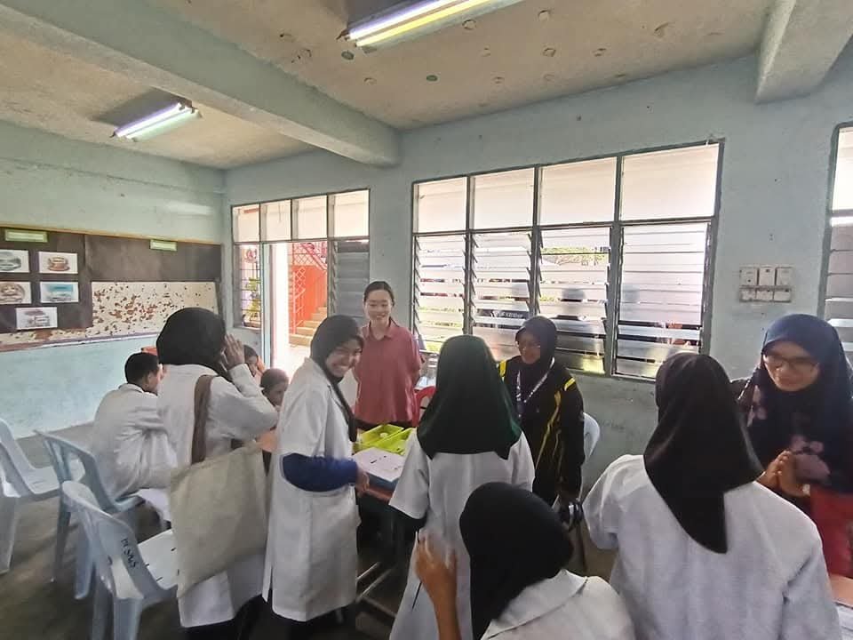 A classroom with students and teachers engaging in a discussion, some students wearing white uniforms and headscarves, in a room with large windows and classroom posters.