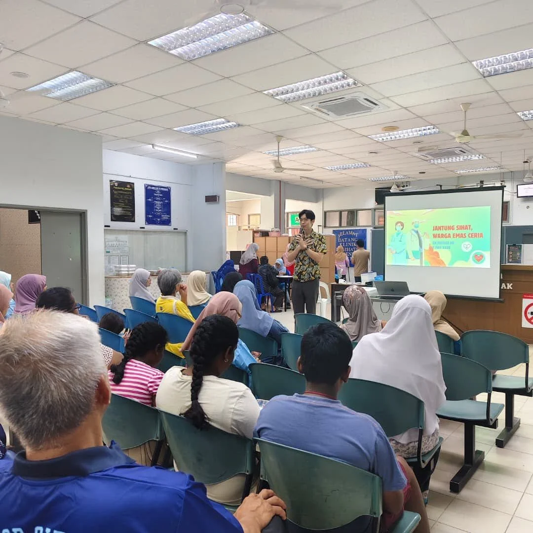 A health seminar in a hospital or clinic setting, with a male presenter speaking to an audience of diverse adults, including women wearing headscarves. A large screen displays a health-related presentation with text. The room has ceiling lights, ceiling fans, and a visible air conditioning unit.