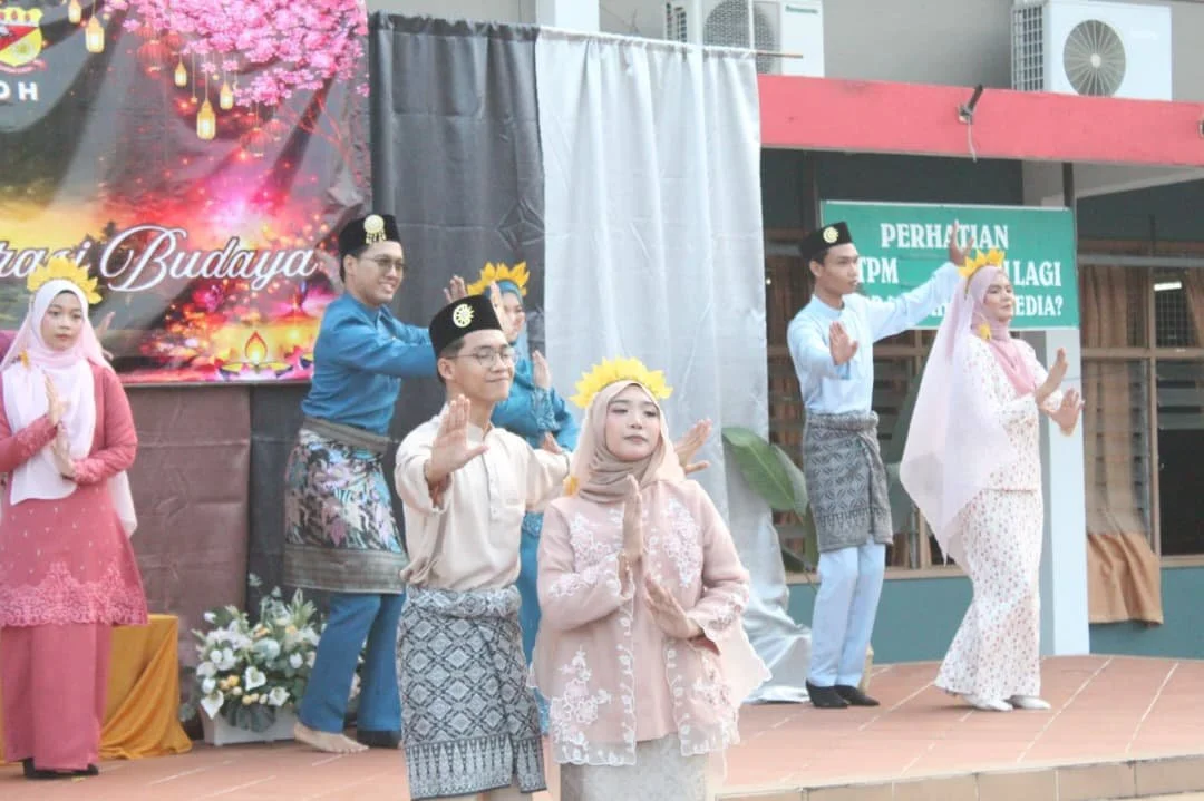 Group of young people dressed in traditional Malay clothing performing a cultural dance on stage, with some wearing yellow flower crowns, in front of a decorated backdrop.