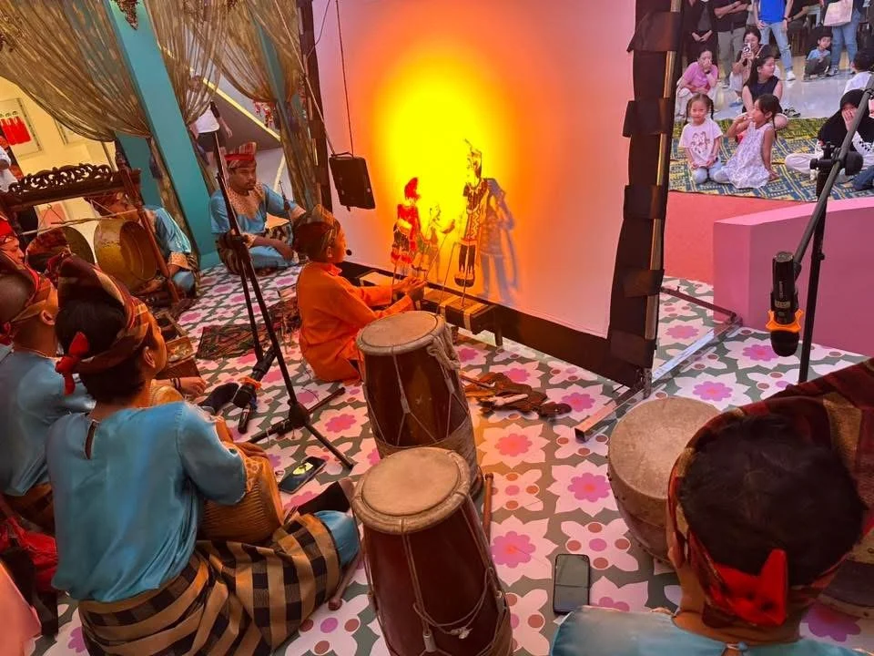 A traditional shadow puppet show with performers and audience, featuring a backdrop with shadow puppets, traditional drums, and children watching in an indoor setting.