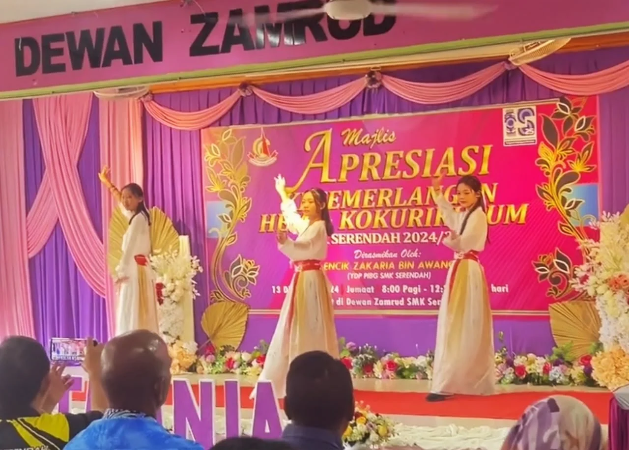 Three girls performing a dance on stage decorated with purple and pink curtains, floral arrangements, and a large pink banner with yellow text that reads 'Majlis Apresiasi Gemilang HEHE Kokurikulum Serendah 2024'. The girls are dressed in traditional
