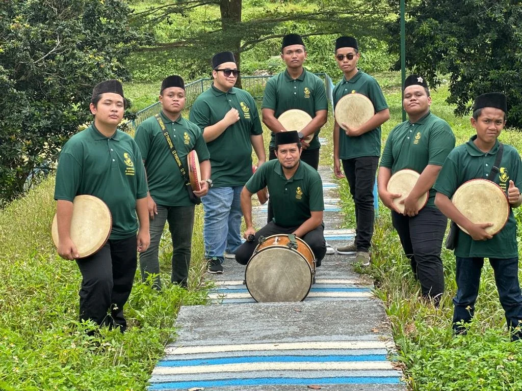 Group of nine boys and one man outdoors on a pathway, holding various percussion instruments, wearing matching green shirts and traditional black hats, with greenery and trees in the background.