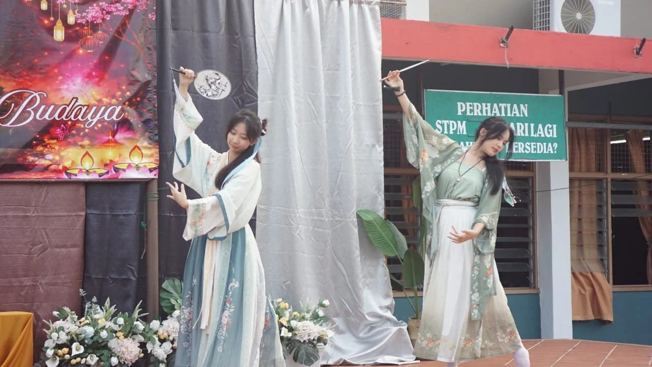 Two women dressed in traditional costumes performing a dance on a stage decorated with flowers, with a colorful backdrop and green sign in Indonesian in the background.