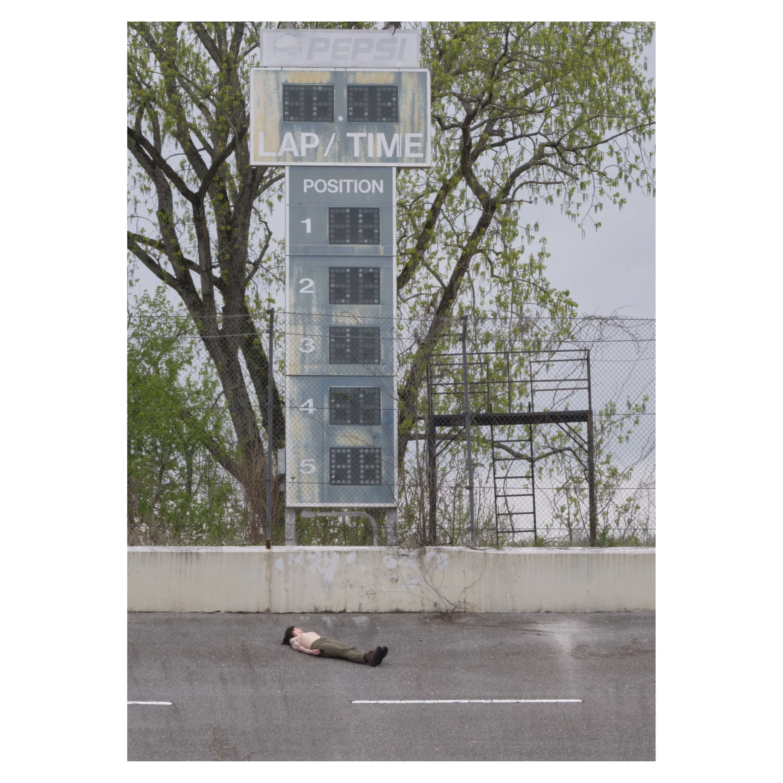 A person lying on the road in front of a large park race scoreboard with trees and chain-link fencing in the background. The scoreboard shows wear to the paint and clearly has not been active in many years.