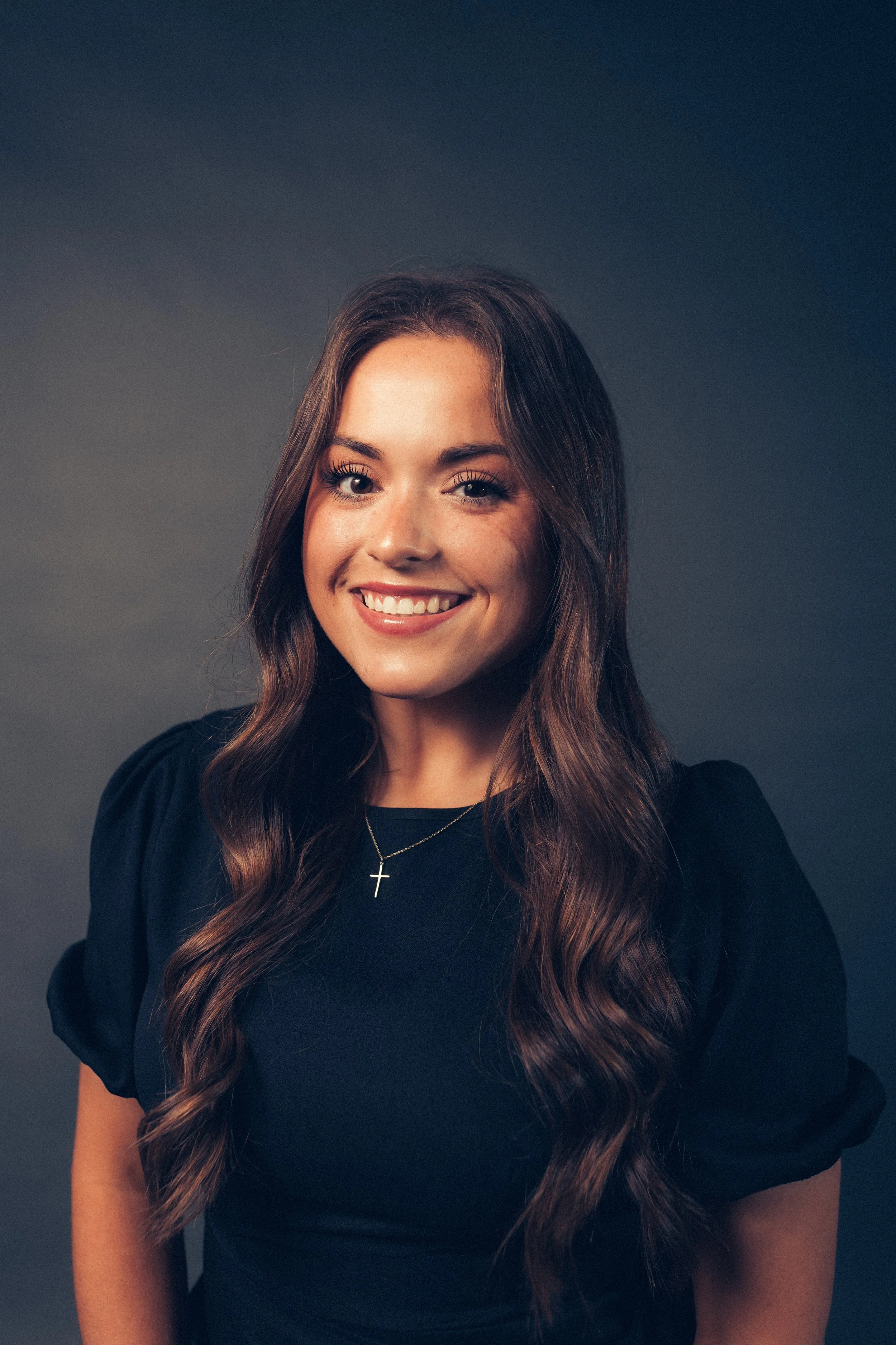 A photo of Reese Sesser, a young woman with long wavy brown hair smiling, wearing a black top and a cross necklace, against a dark background.