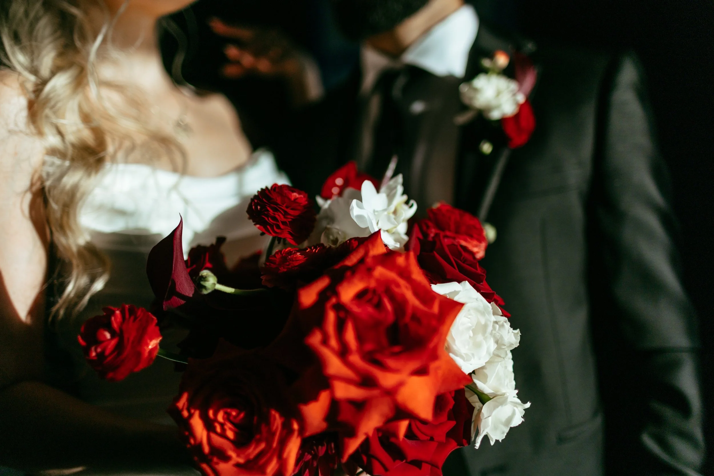 Close-up of a bride holding a bouquet of red, white, and dark pink flowers at a wedding.