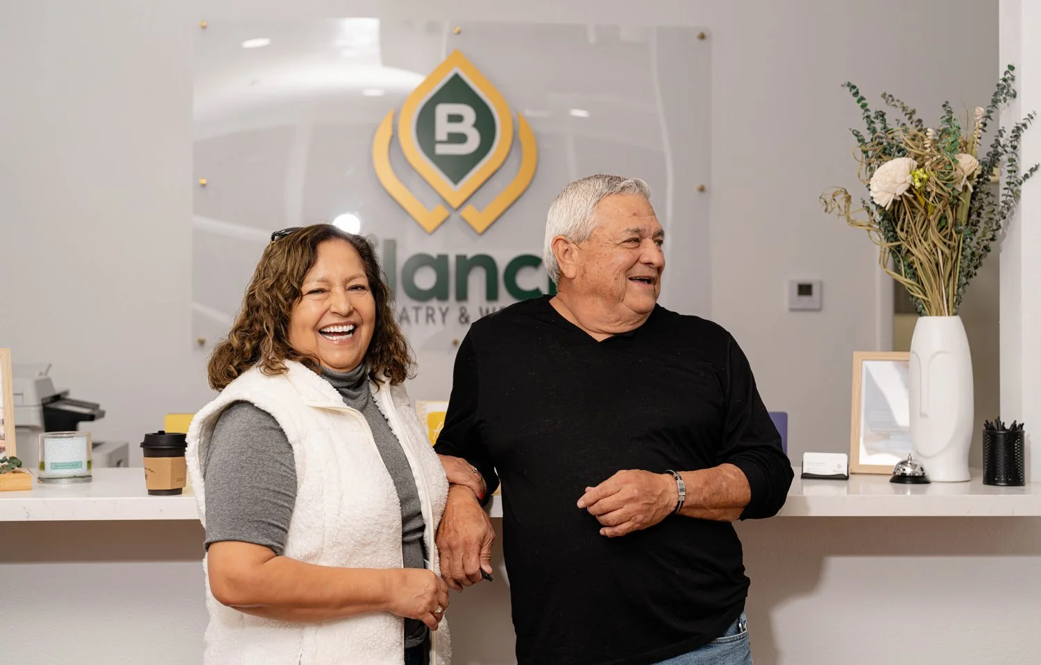 Two happy people, a woman and a man, standing at a reception desk in an office, smiling and holding hands, with a logo behind them reading "Lance".