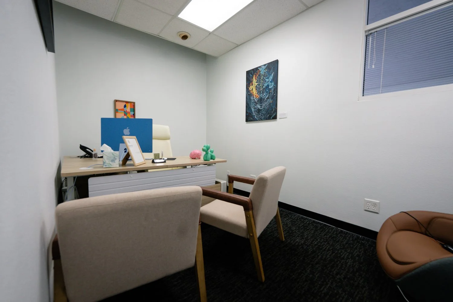 Empty office with two chairs in front of a desk holding a Mac monitor, tissue box, framed picture, and balloons, with a window on the right and colorful artwork on the walls.