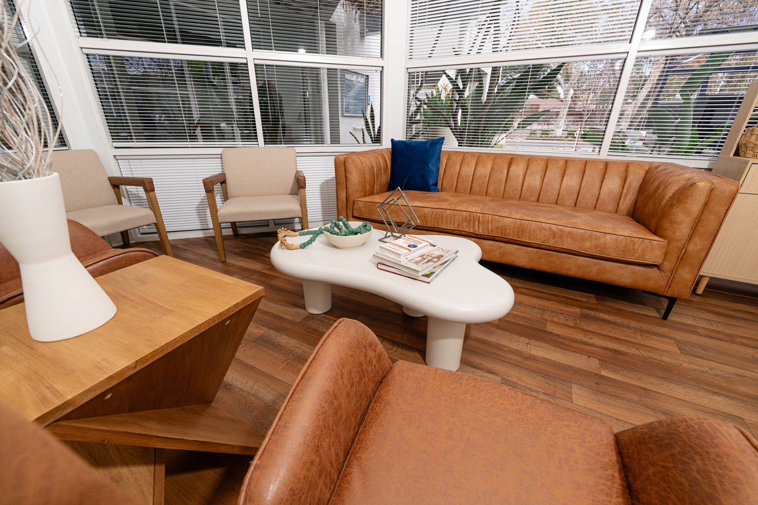 A cozy waiting room with beige chairs, a tan leather sofa with a blue throw pillow, and a white curvy coffee table with books and decorative items, overlooking large windows with blinds revealing a plant outside