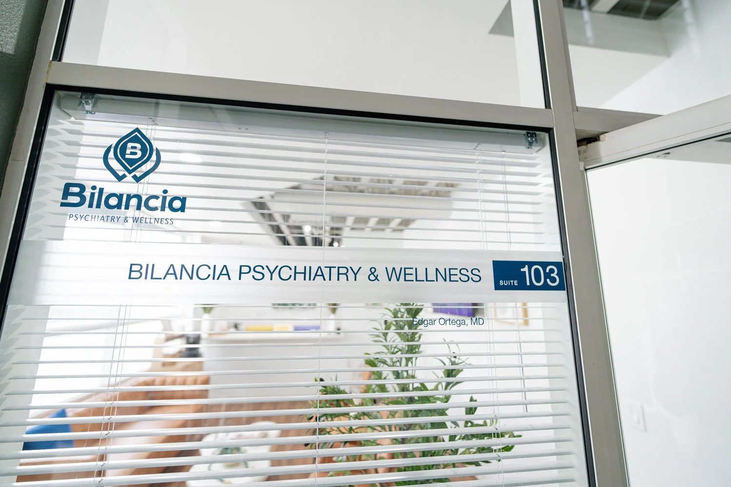 Glass door office window with blinds showing signs for Bilancia Psychiatry & Wellness, Suite 103, and Dr. Edgar Ortega, MD, with a glimpse of an office interior and a potted plant inside.