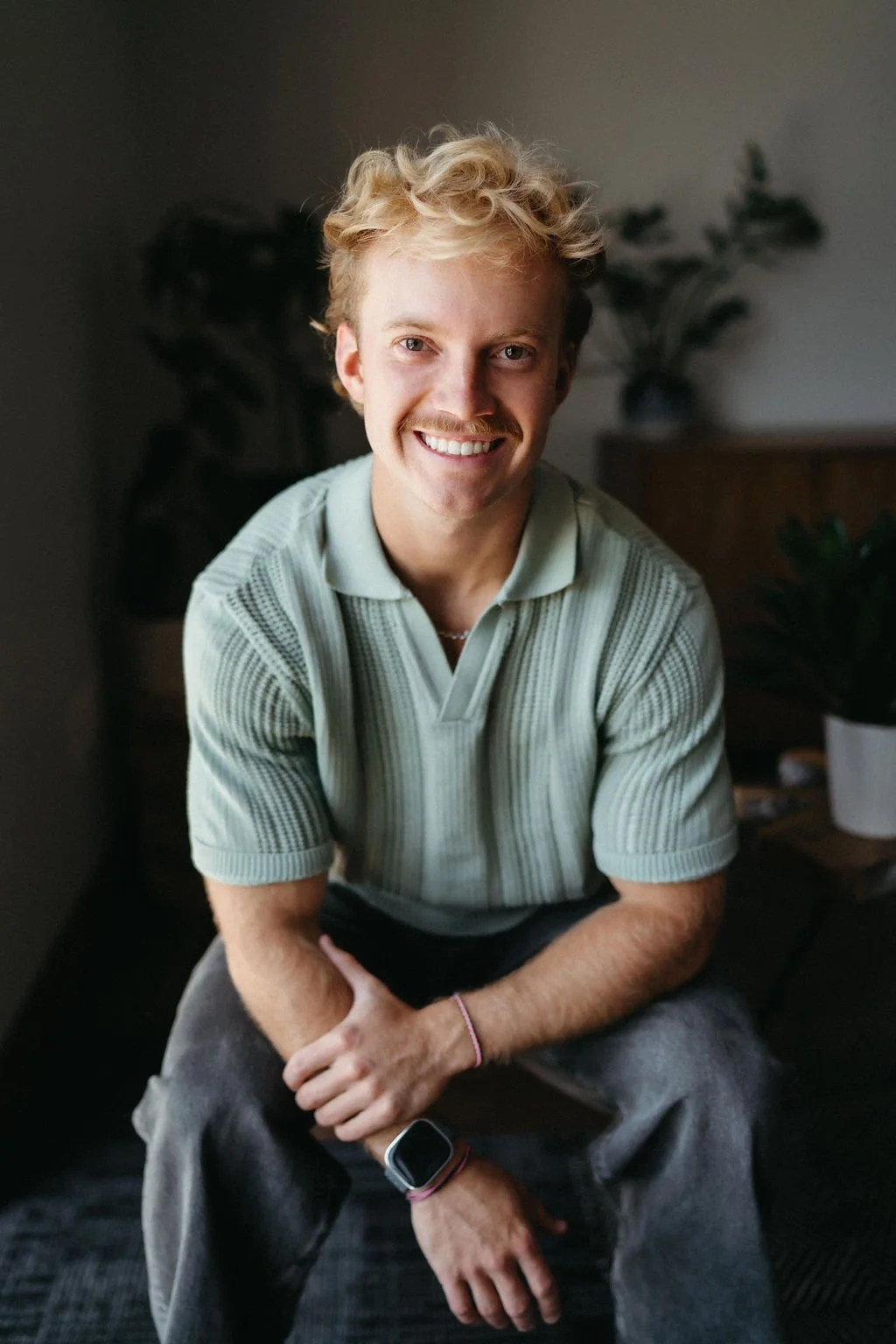Young man with blond curly hair smiling outdoors in a rural setting, wearing a black Nike sweatshirt and silver chain necklace.