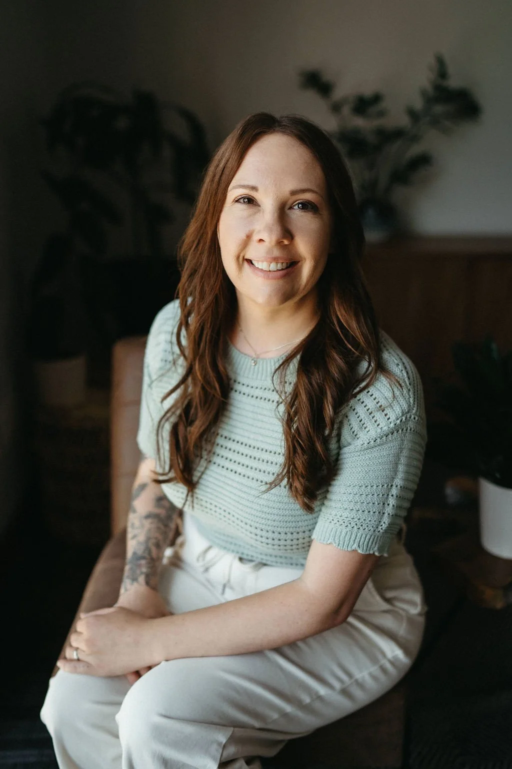 A woman with long brown hair and a mustard yellow top smiling indoors, with a background featuring a bookshelf, plants, and a landscape painting.