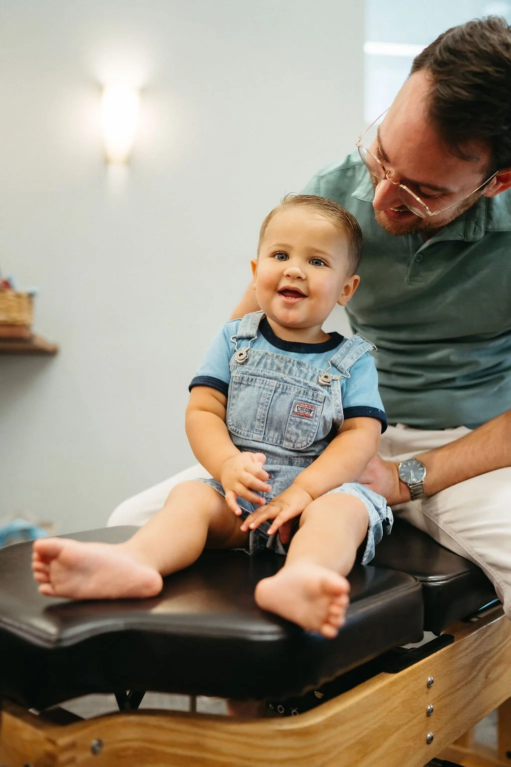 A baby with spiky hair lying on a blue bandana-patterned pillow on a black chiropractic adjustment table, being gently held by two hands.