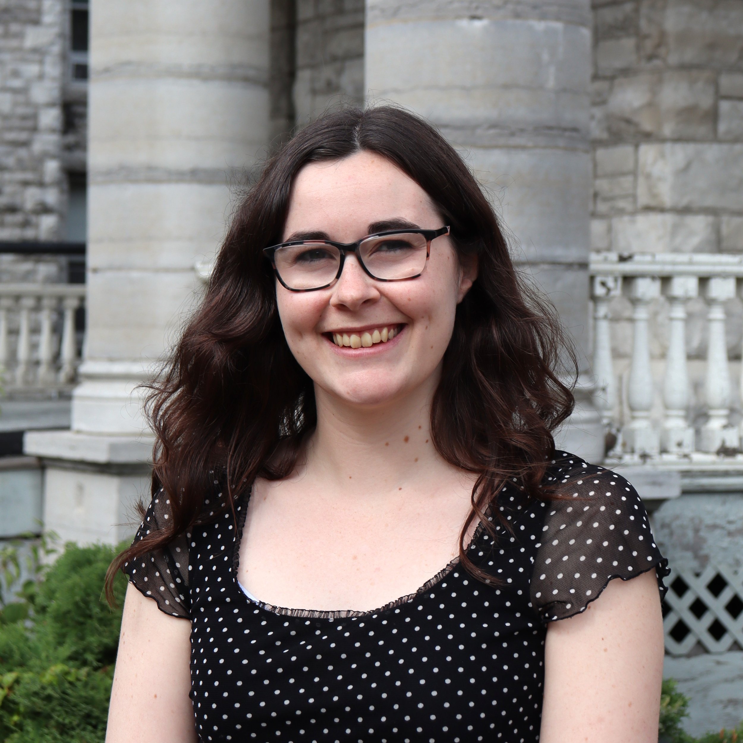 A young woman with brown hair, glasses, and a black polka dot shirt smiling in front of a stone wall.