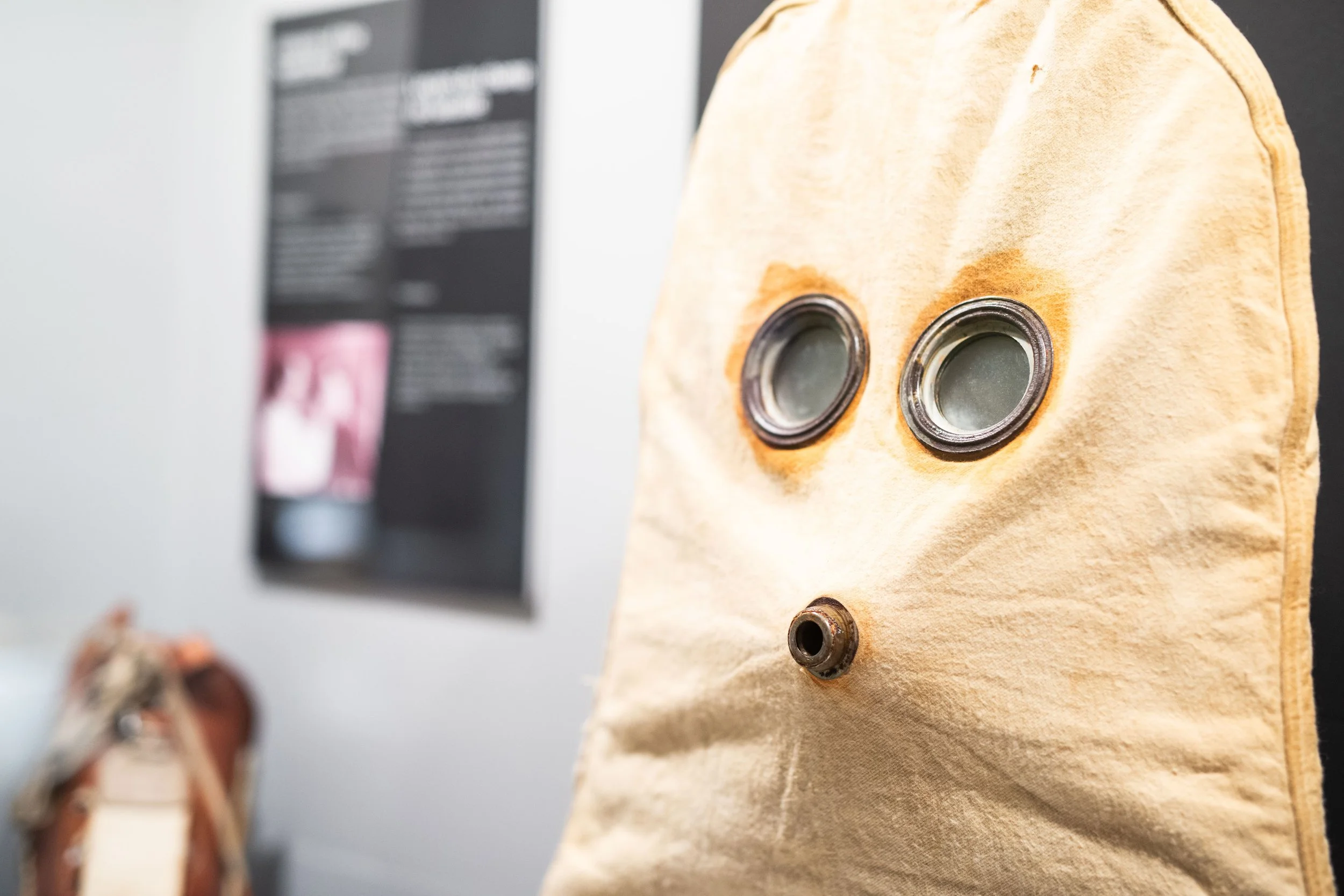 A beige fabric bag with two metal and glass valves and one metal opening, possibly a protective mask or filter, standing upright in front of a blurred black and white informational poster.