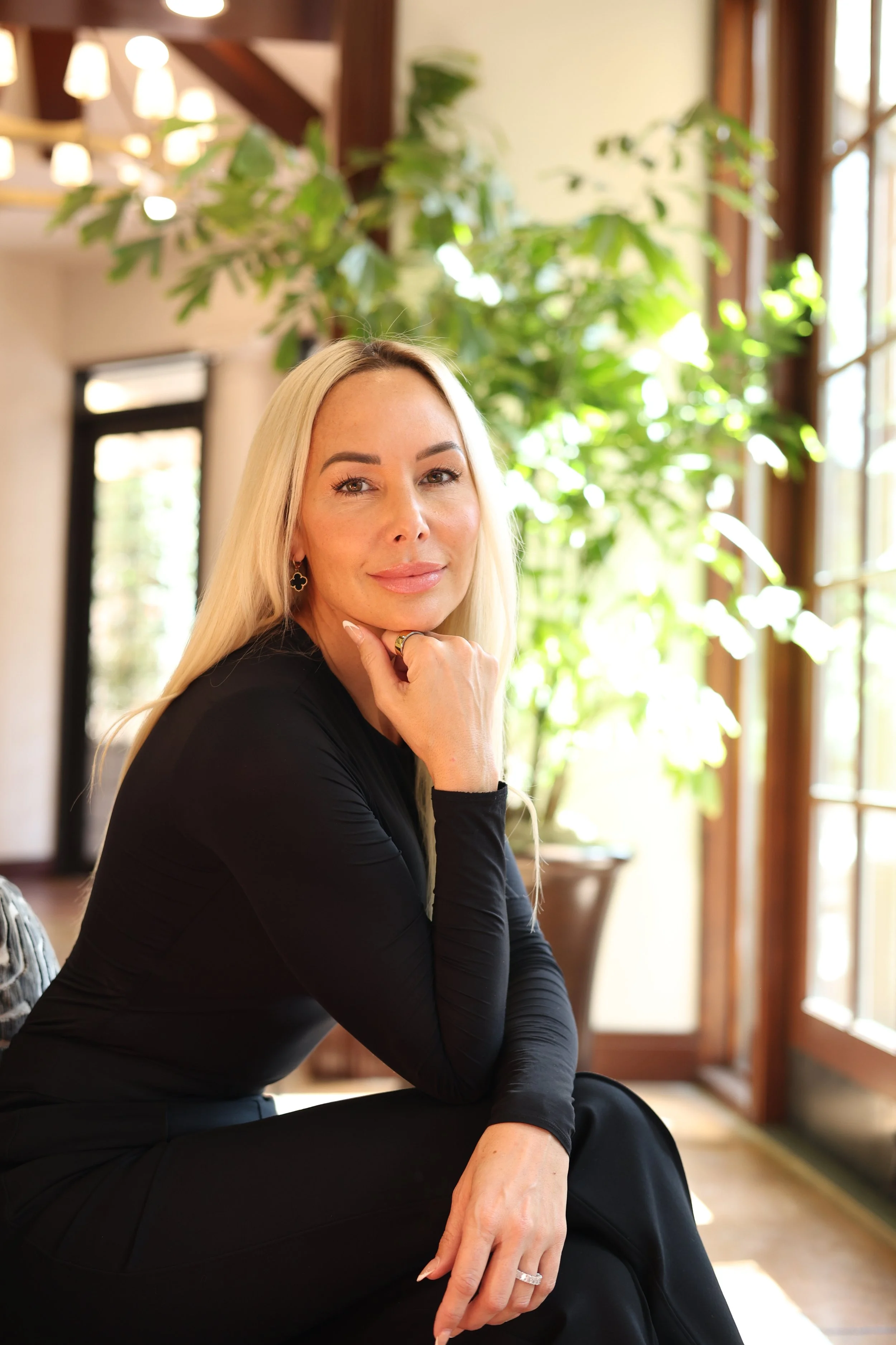 A woman with long blonde hair, wearing black clothing, sitting indoors near large windows with sunlight and greenery in the background.