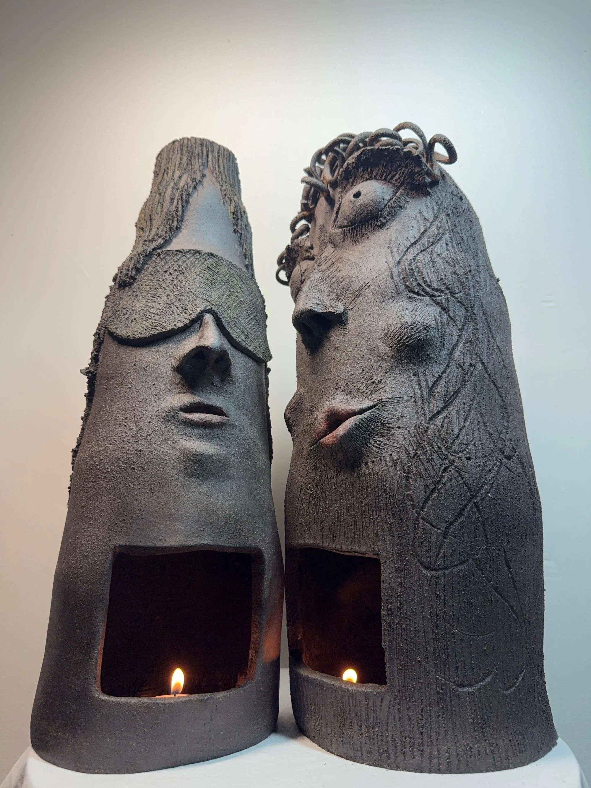 Two black stoneware ceramic sculptures titled 'Dude and Dudette', featuring expressive chiseled facial features, hand-coiled hair curls, and a functional candle chamber at the base.
