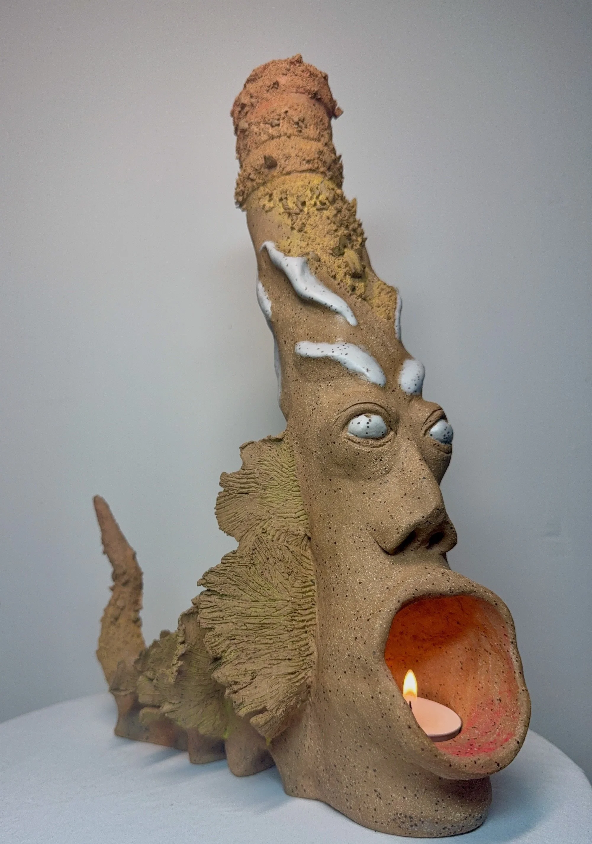 A warm toned stoneware ceramic sculpture titled: ‘Rebirth of a Sunny Day’, featuring a dragon-like shape with wings and pointed tail, a functional candle chamber at the base, and a tall turban that allows for smoke to come out of the top.