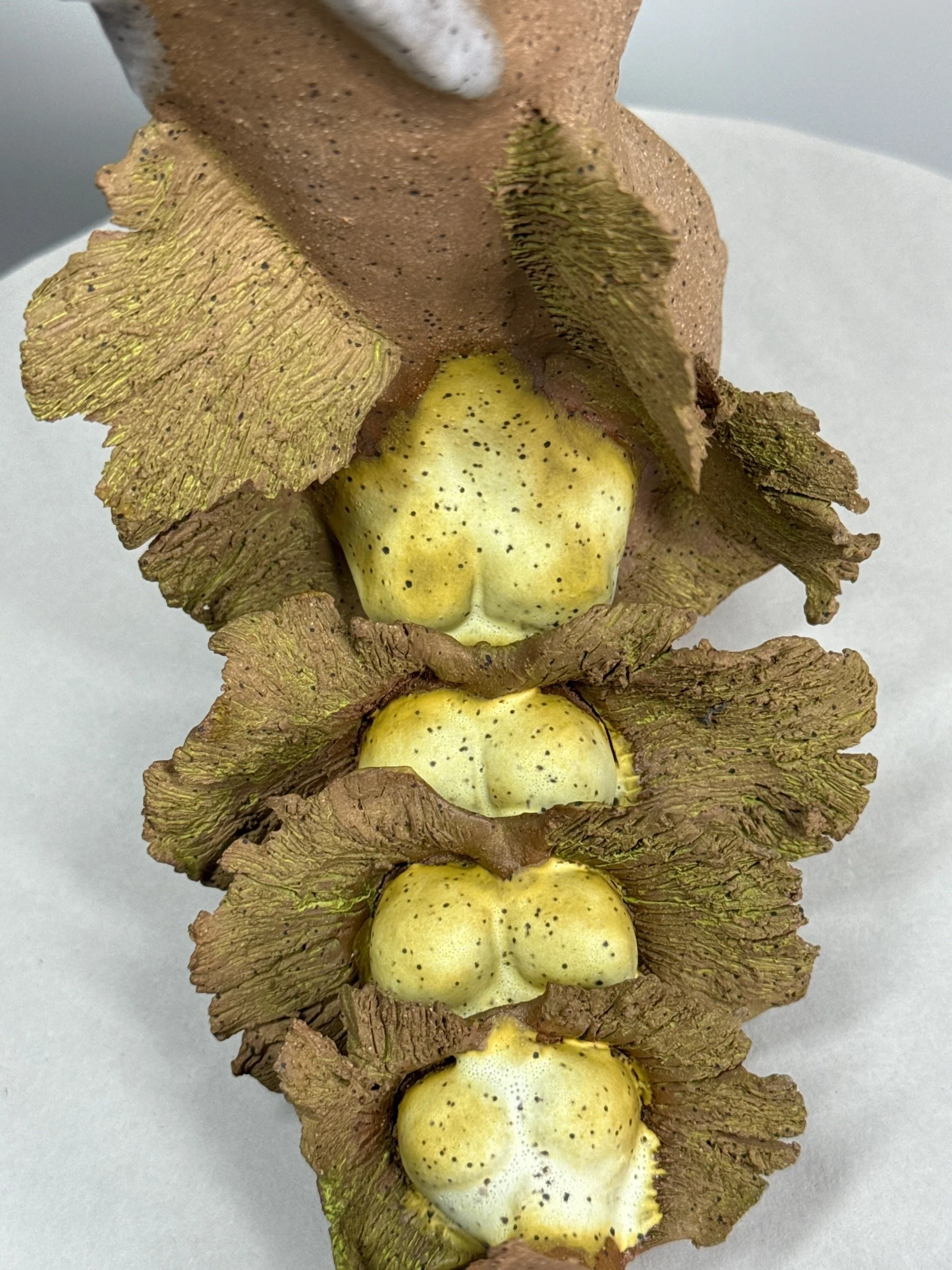 A detail of a warm toned stoneware ceramic sculpture titled: ‘Rebirth of a Sunny Day’, featuring a dragon-like shape with dramatically carved wings and bulbous backbones in yellow.