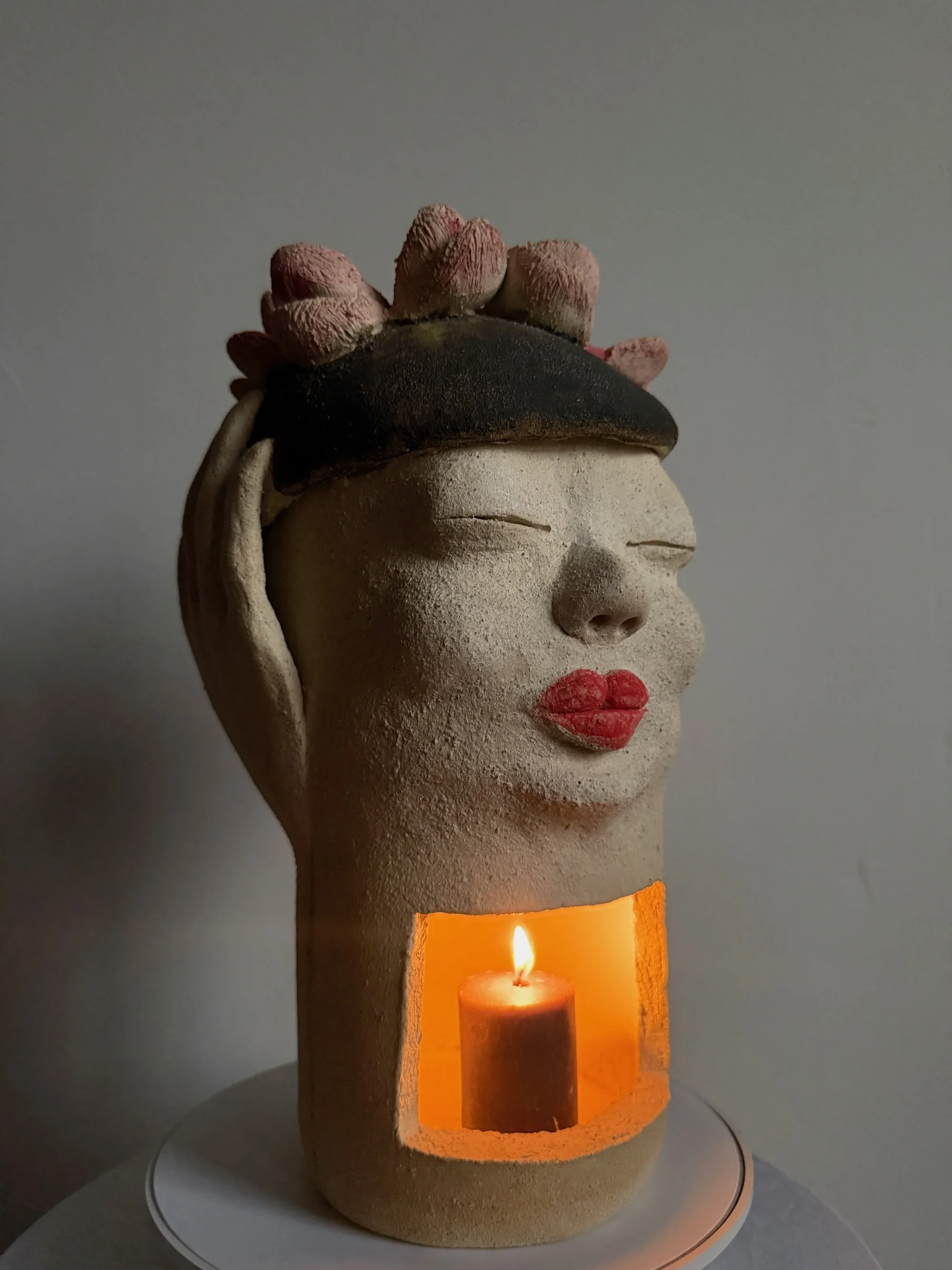 A white stoneware ceramic sculpture titled ‘Beret Adjustment’, featuring full red lips, a black beret with pink flowers on top, and a functional candle chamber at the base.