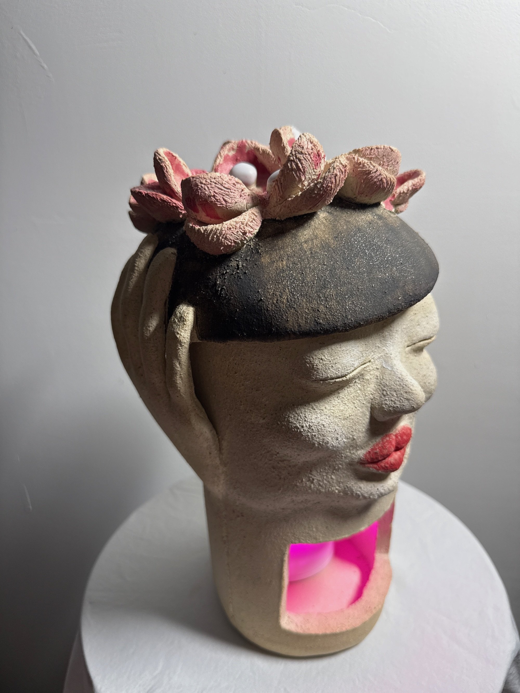 A white stoneware ceramic sculpture titled ‘Beret Adjustment’, featuring full red lips, a black beret with pink flowers on top, and a pink LED light in the base