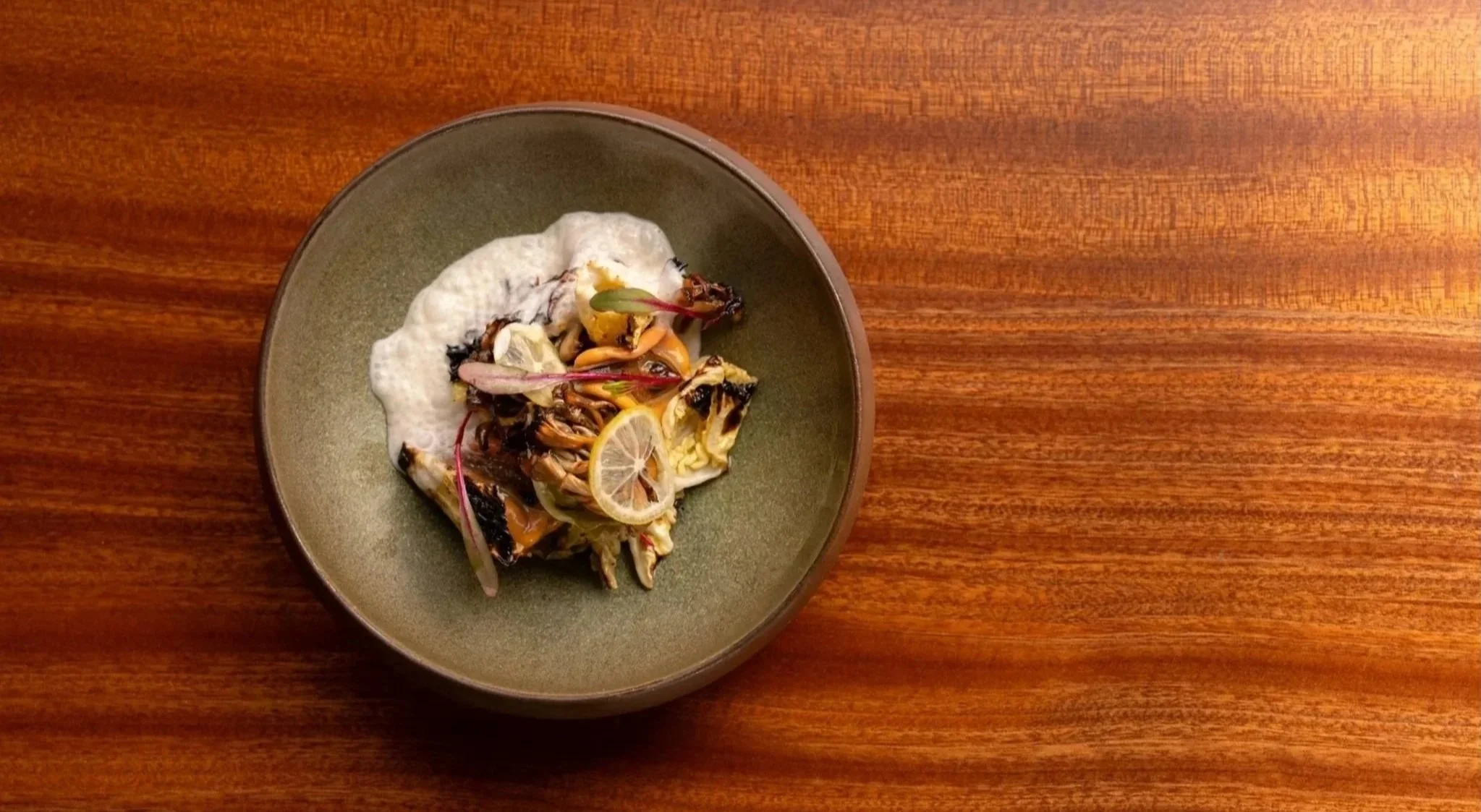 A green ceramic bowl containing maitake, mussels, brassica, horseradish emulsion garnished with lemon slices and microgreens, served on a wooden surface. 