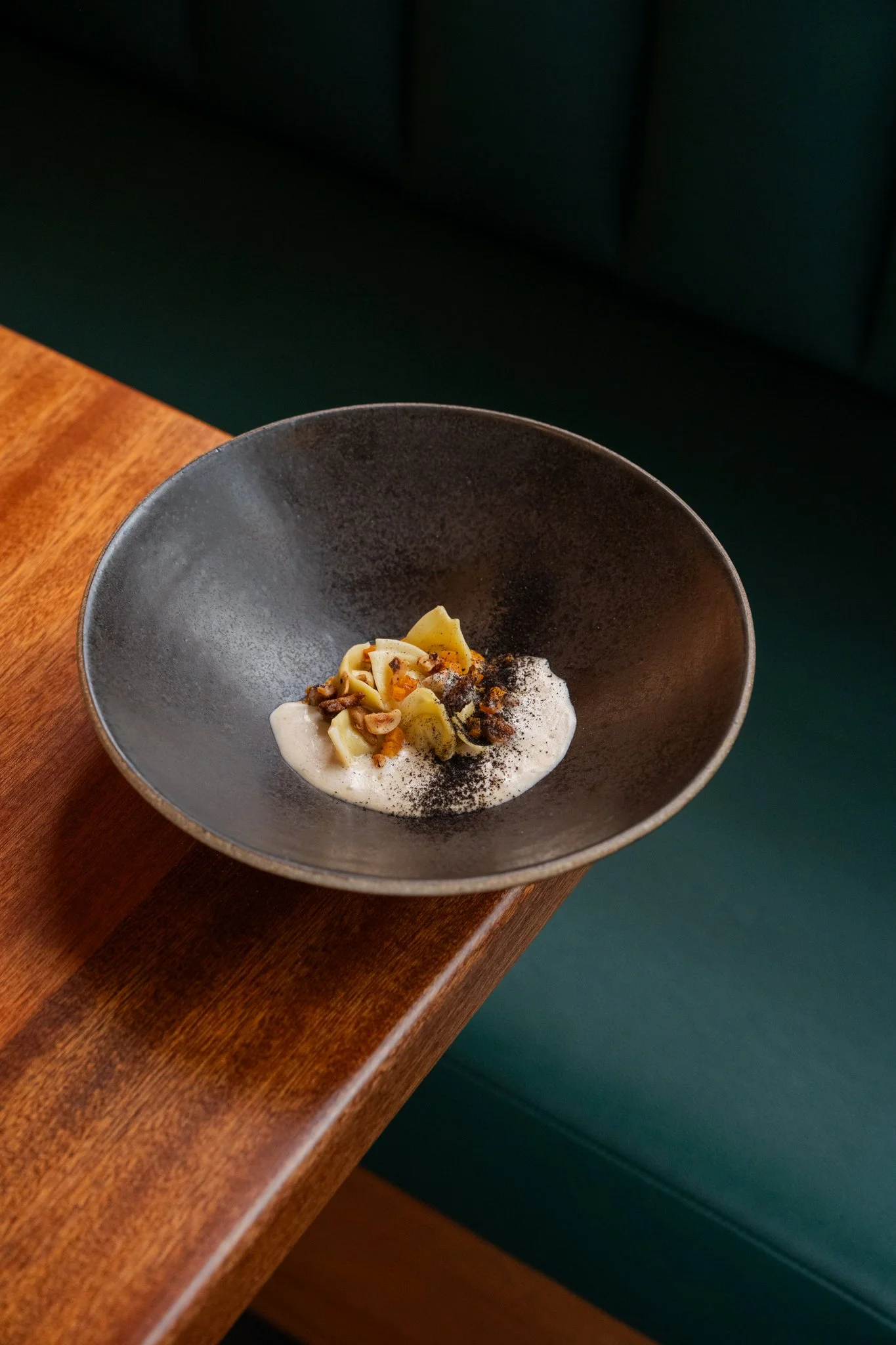 A black ceramic bowl with a serving of cappellaci pasta with butternut squash, brown butter topped with hazelnuts and ash of allium set on a wooden table.
