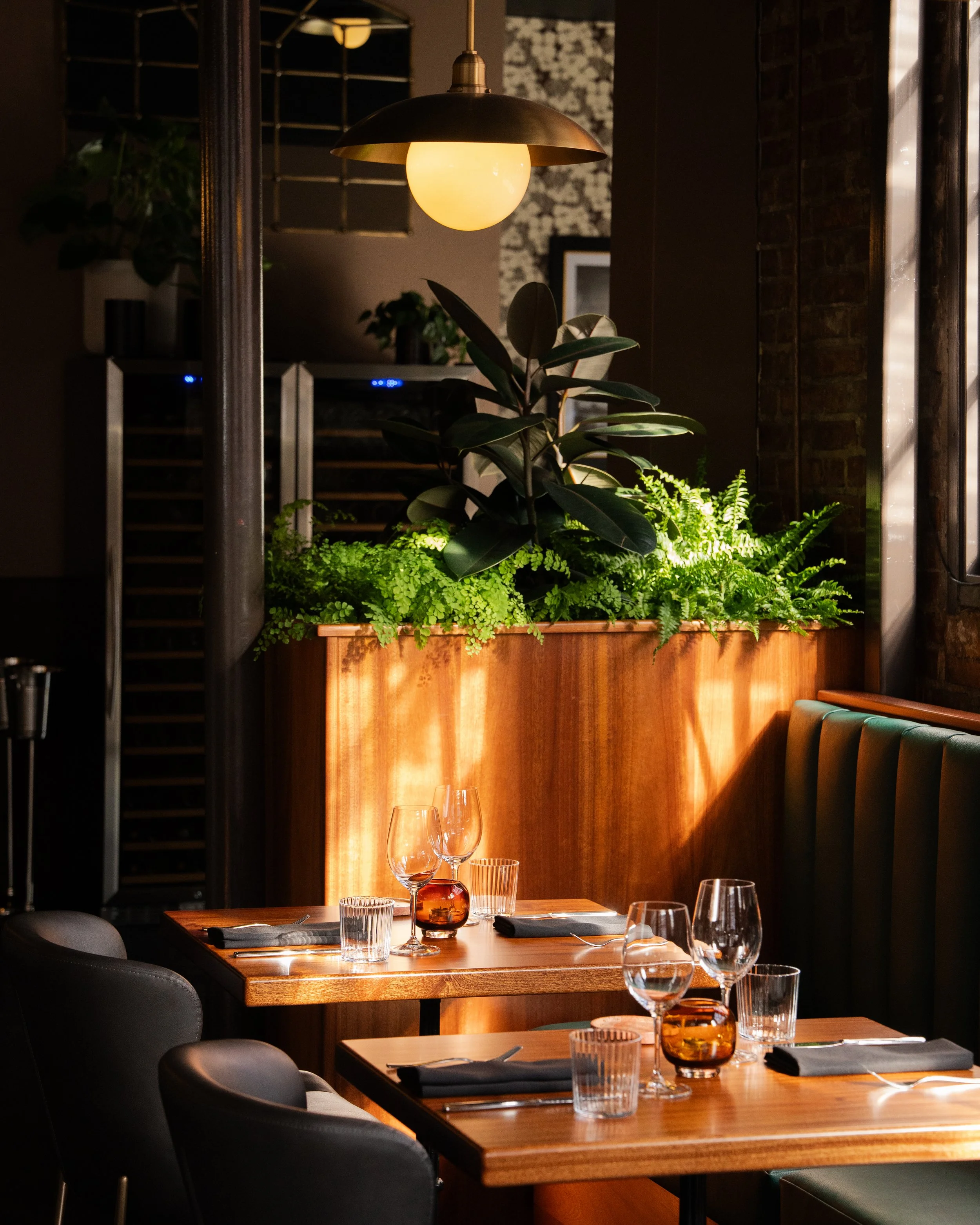 Restaurant interior with set tables with green benches, wooden accents, lush plants and moody lighting.