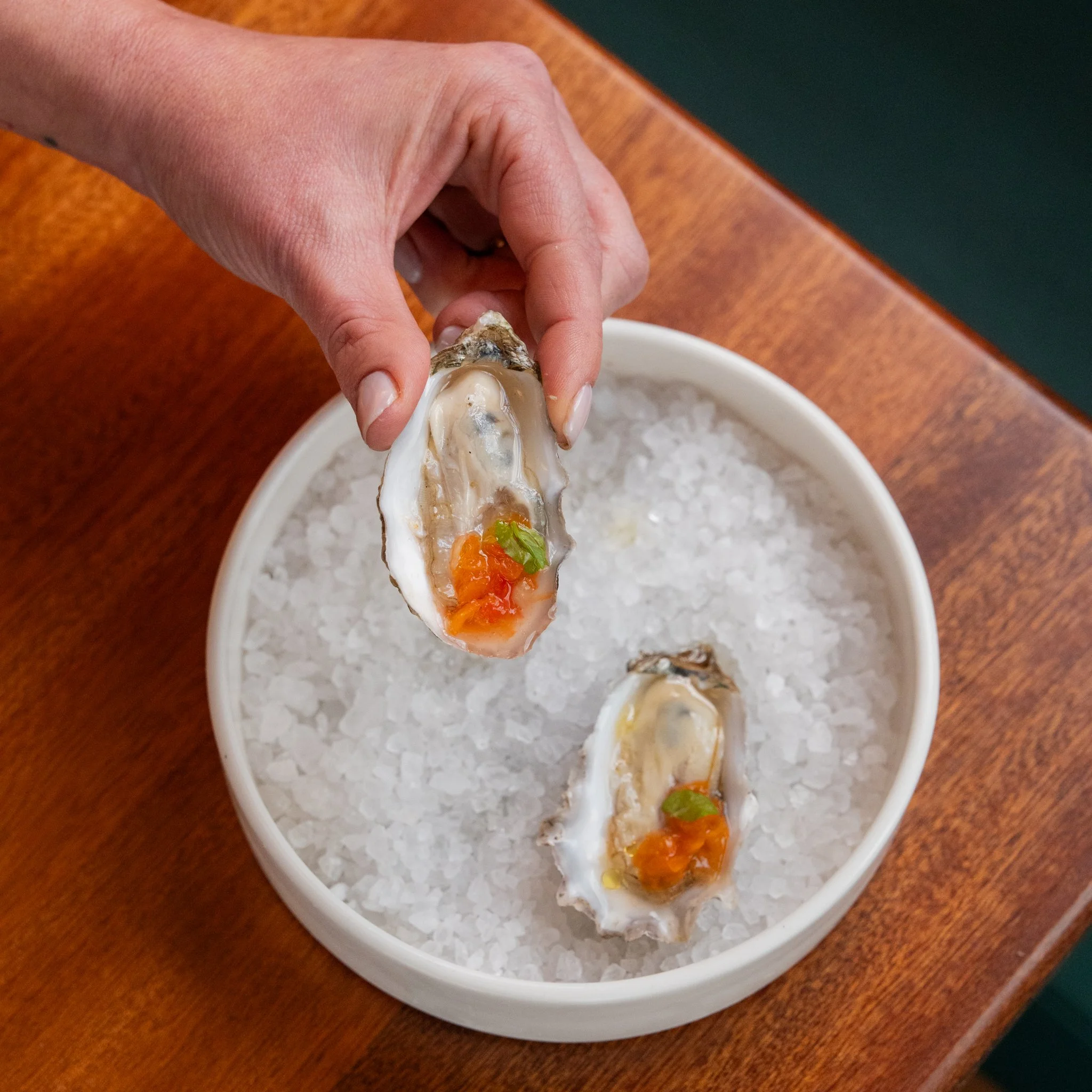 A hand holding an oyster with orange and green garnish, over a bowl of crushed ice containing another oyster with similar garnish.