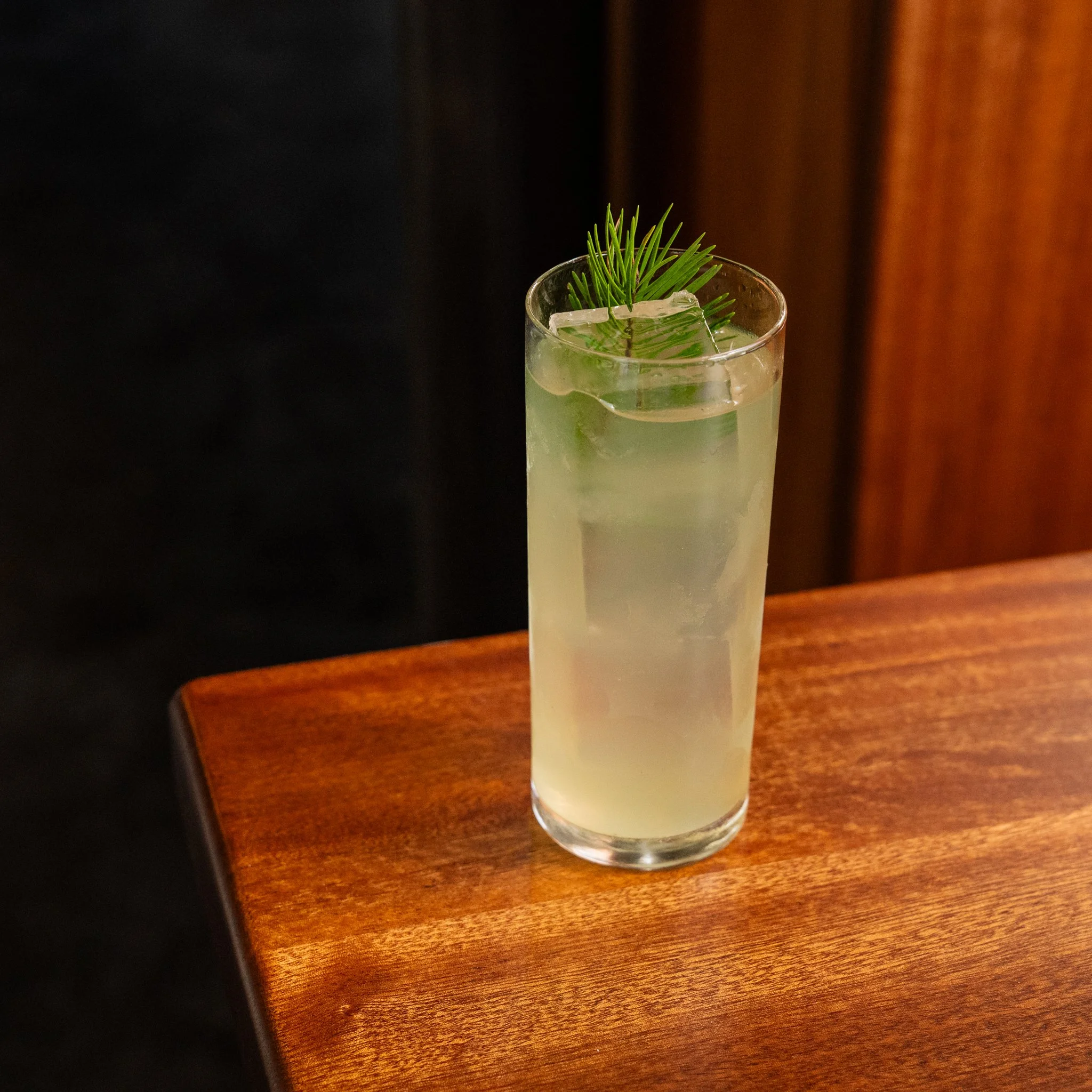 A highball glass with a pale yellow-green cocktail, ice cubes, and a garnish of douglas fir, on a wooden table.