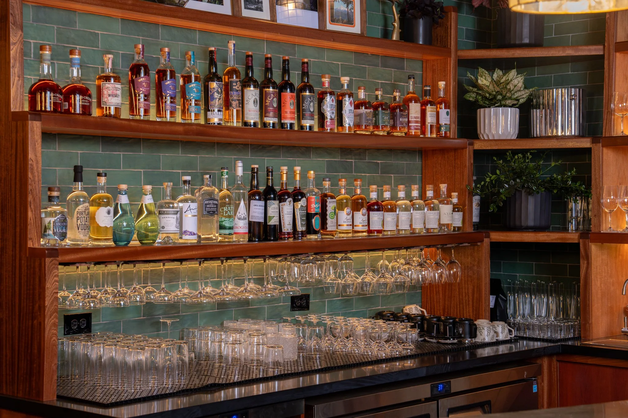 A bar shelf with bottles of liquor, wine, and spirits, along with glassware, wine glasses, and decorative plants.