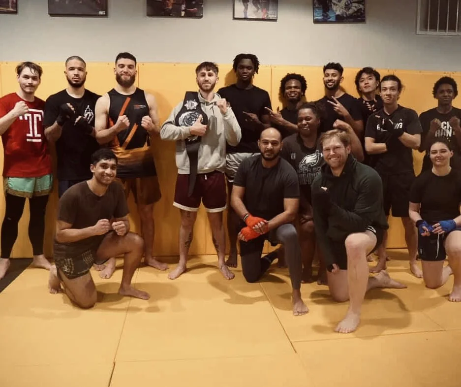 Group of people in a gym posing for a photo, some in martial arts attire, with trainers kneeling in front.