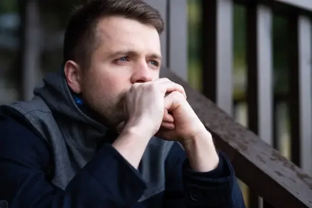 A man with short brown hair and a beard sitting outdoors on a wooden bench, resting his chin on his clasped hands, looking thoughtfully into the distance.
