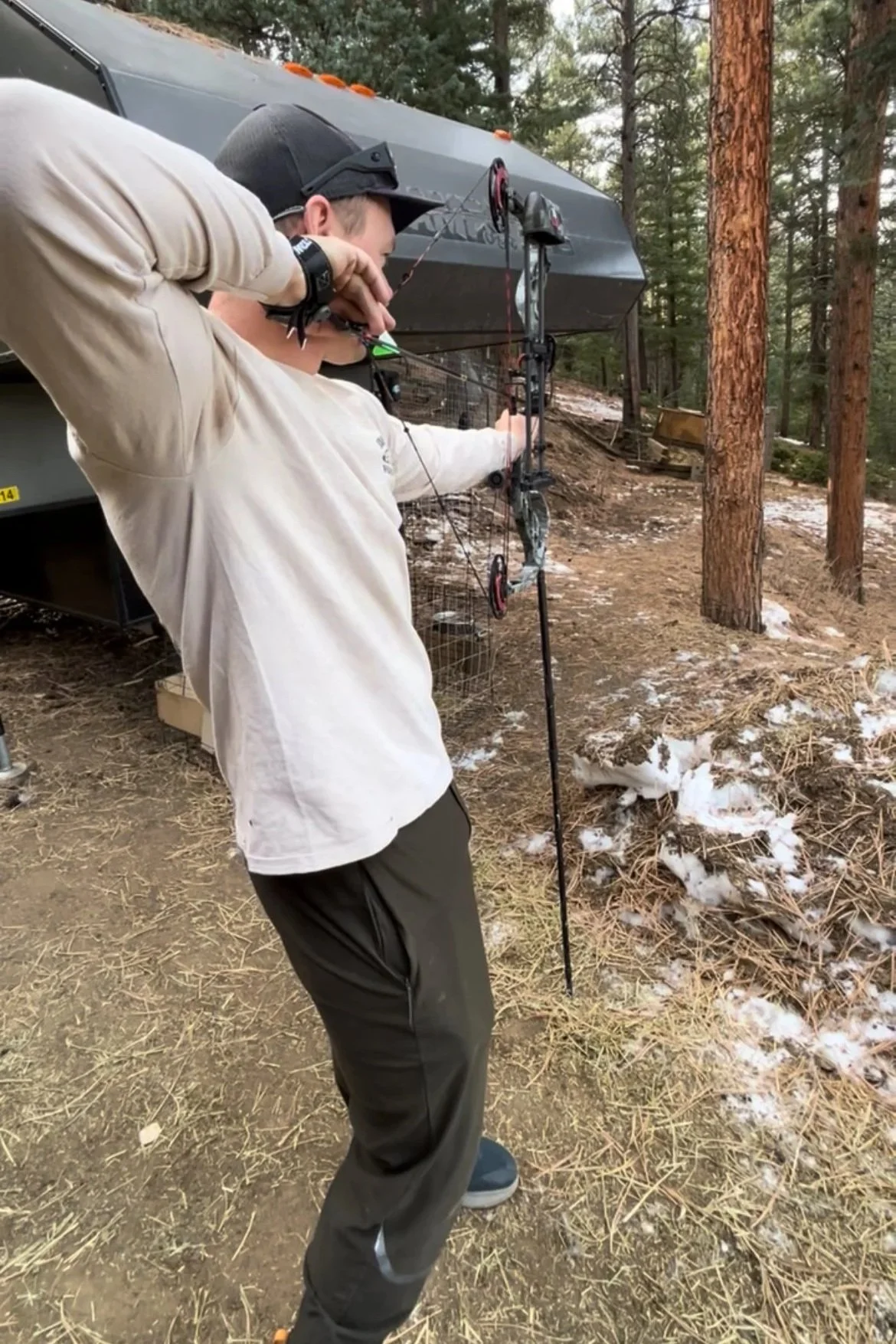 Nick, the founder aiming a bow and arrow outdoors near a camper trailer in a forested area with pine trees and patches of snow on the ground, using the bow paw archery monopod.