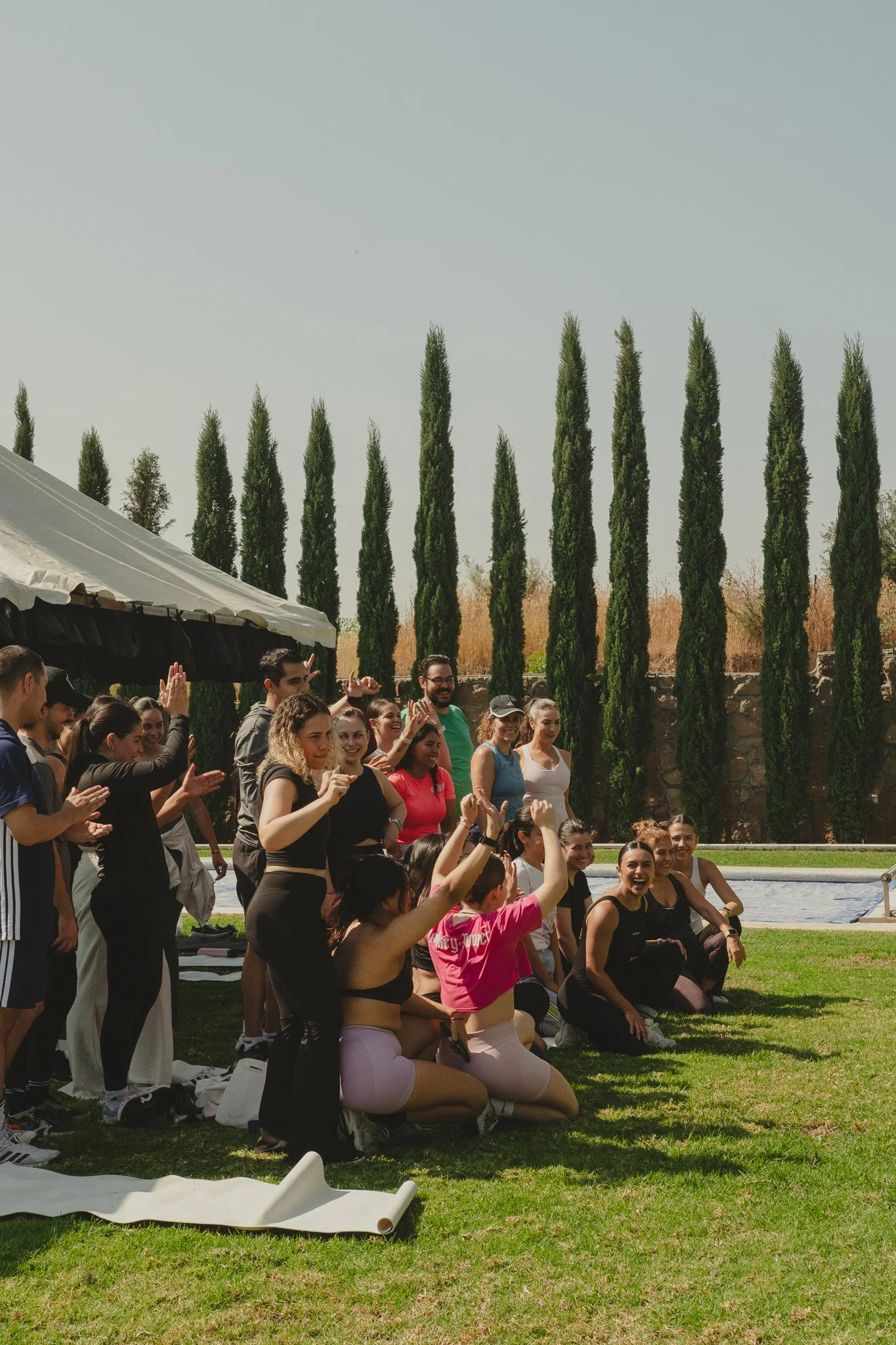Grupo de personas en ejercicio al aire libre, algunos en posición de rodillas, otros de pie, sonriendo y levantando las manos en señal de celebración o saludo, con árboles altos y césped verde en el fondo.