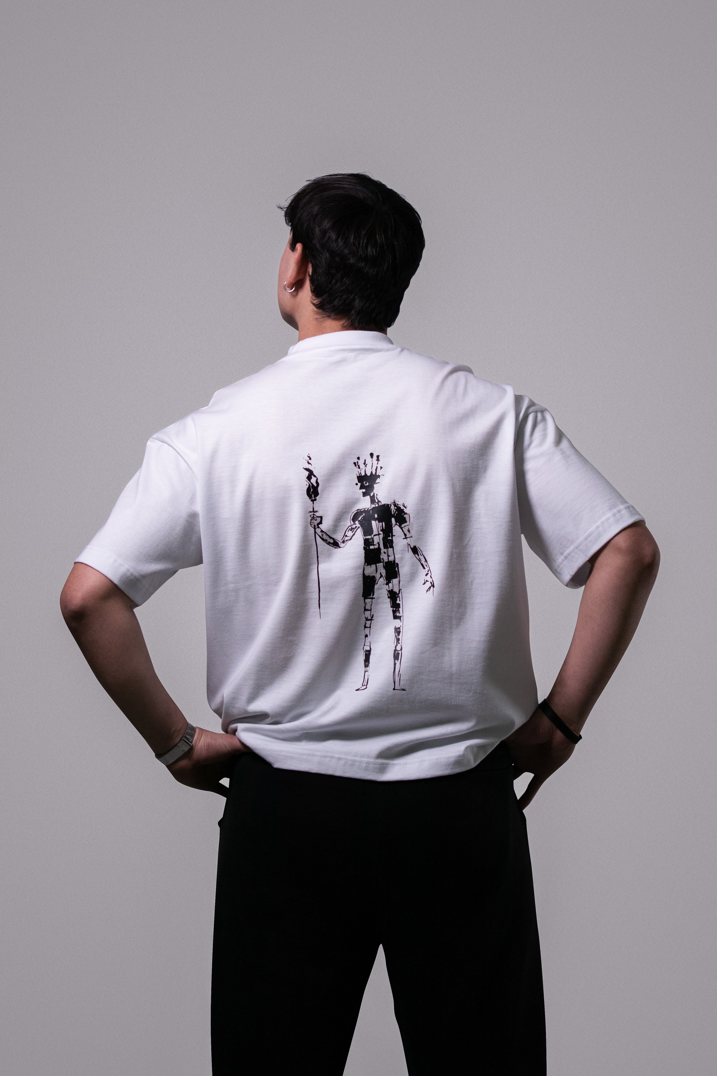 Persona de espaldas con camiseta blanca que tiene un dibujo de una figura humanoide con barba, sosteniendo un tridente, en un fondo gris.