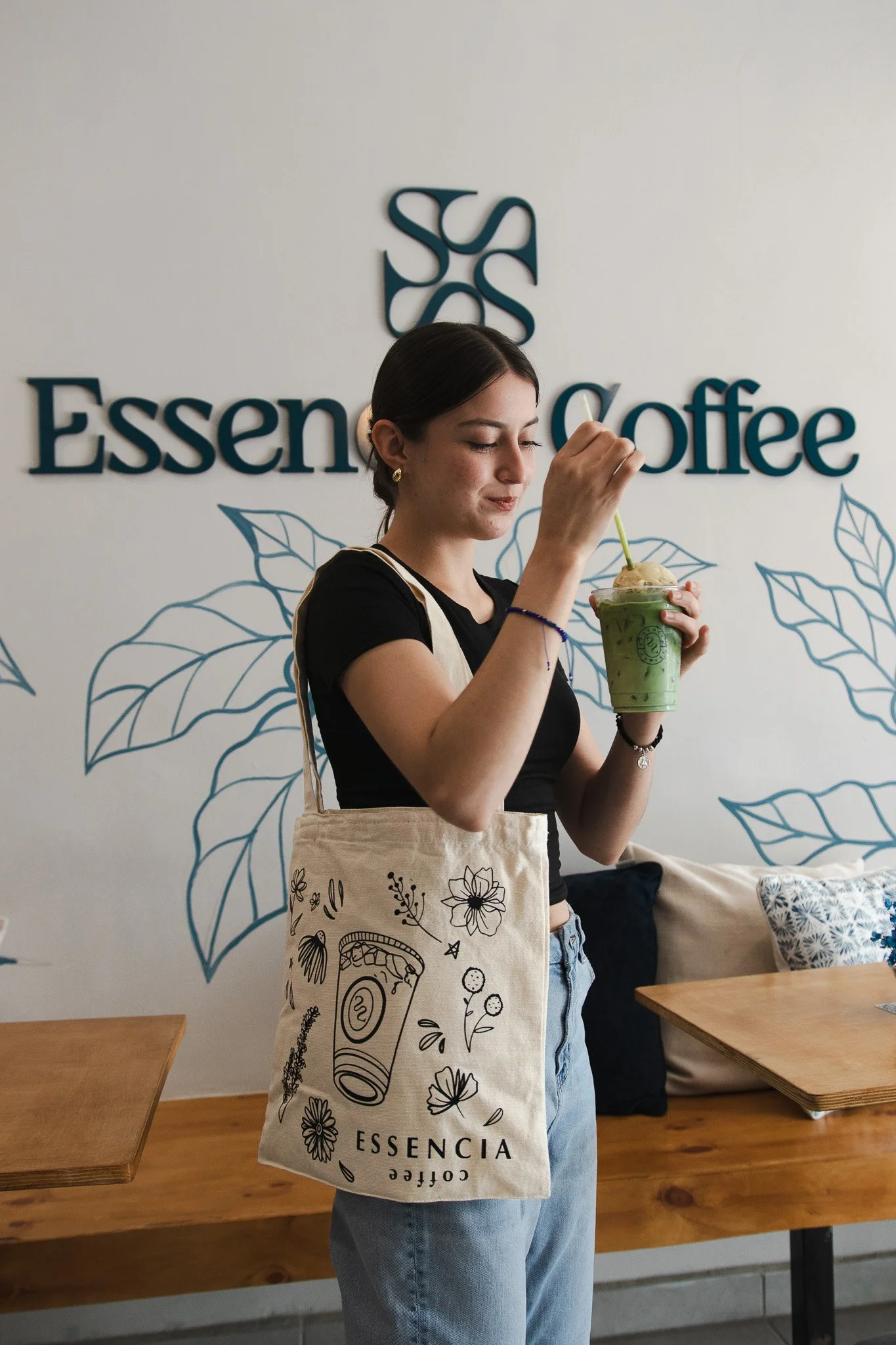 Joven mujer en cafetería sostiene bebida verde en vaso con popote, lleva bolsa de tela con diseño floral y logotipo 'ESSENCIA Coffee'.