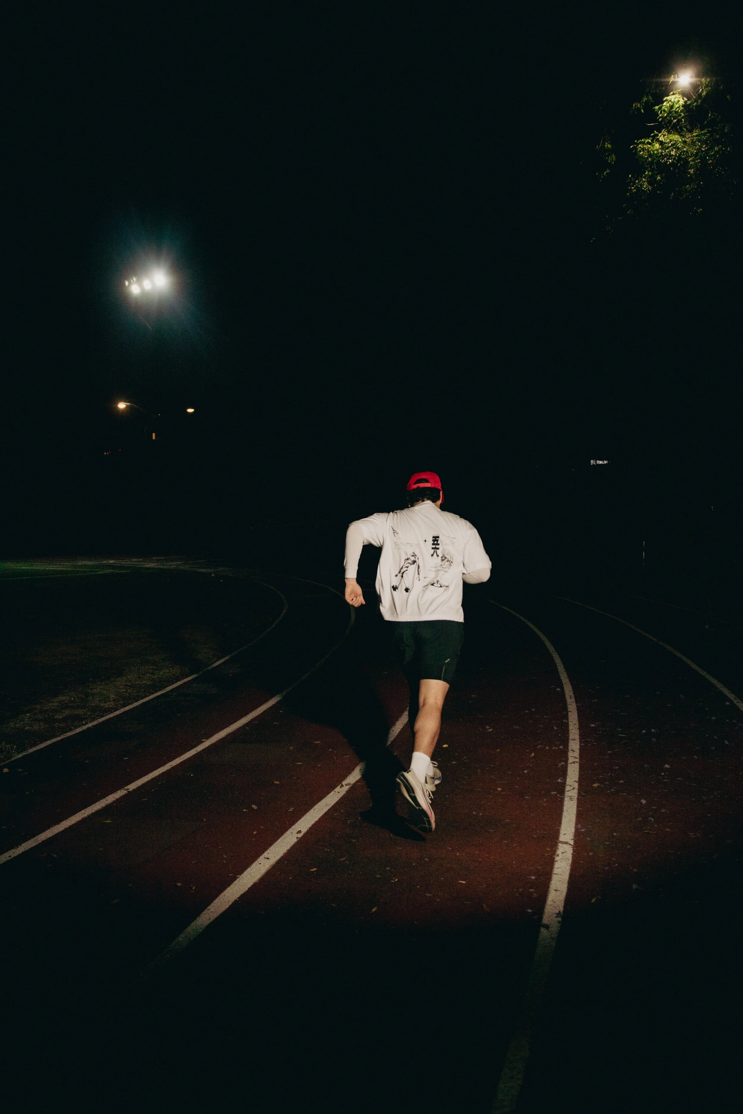 Una persona corriendo en una pista de atletismo de noche, vestida con ropa deportiva blanca y una gorra roja, bajo luces y árboles.