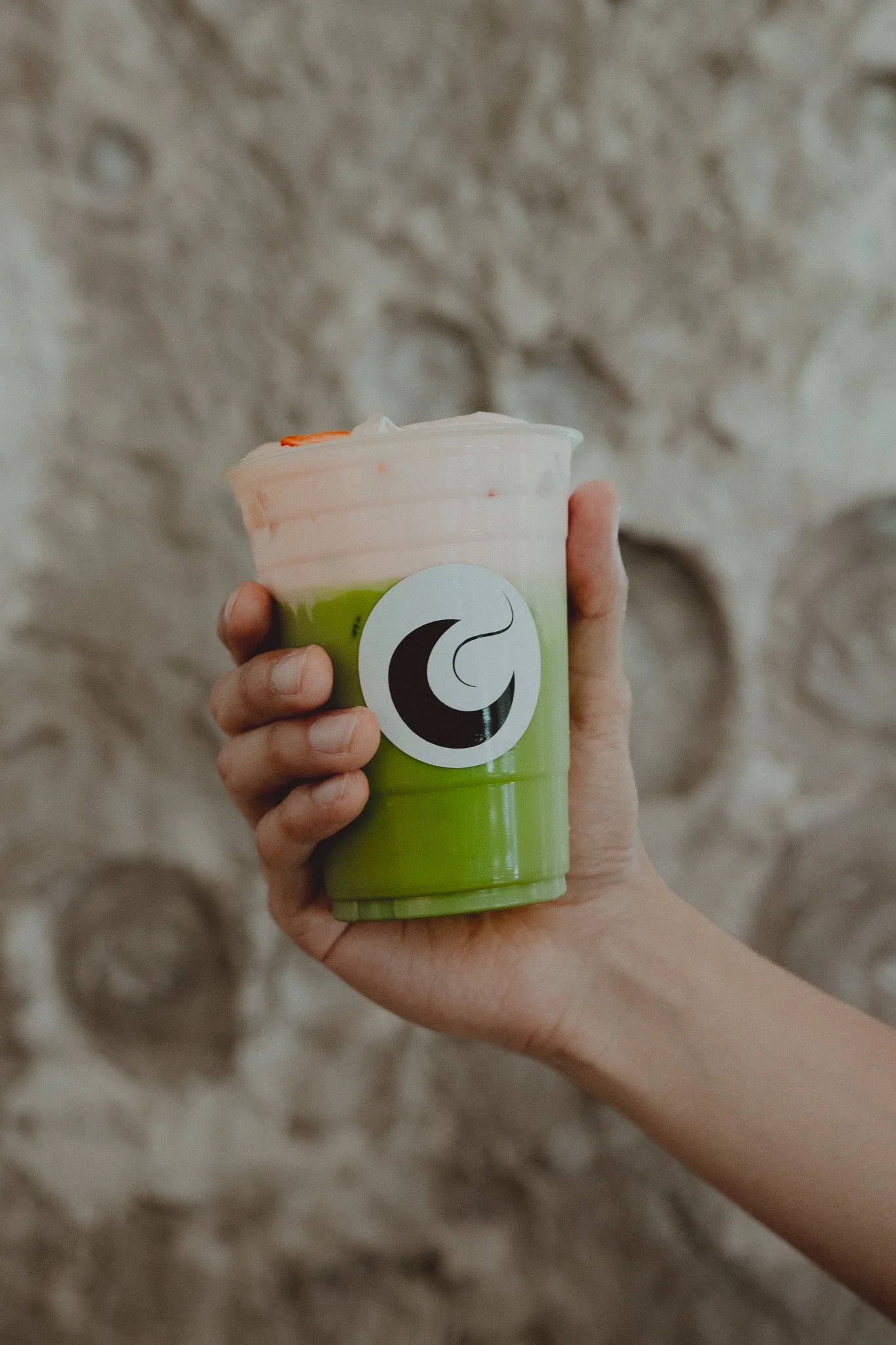 Persona sosteniendo un vaso con una bebida verde en la parte inferior y una espuma rosada en la parte superior, con un logotipo en el centro del vaso, frente a una pared de piedra.