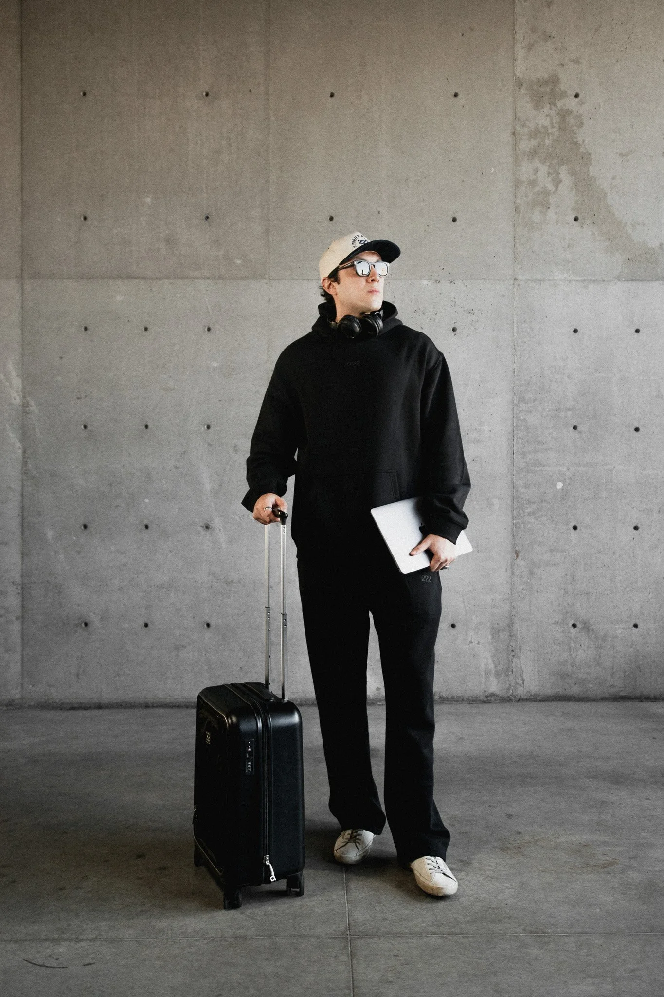 Joven con ropa casual, mochila con ruedas y maletín en mano, en un lugar con paredes de concreto.